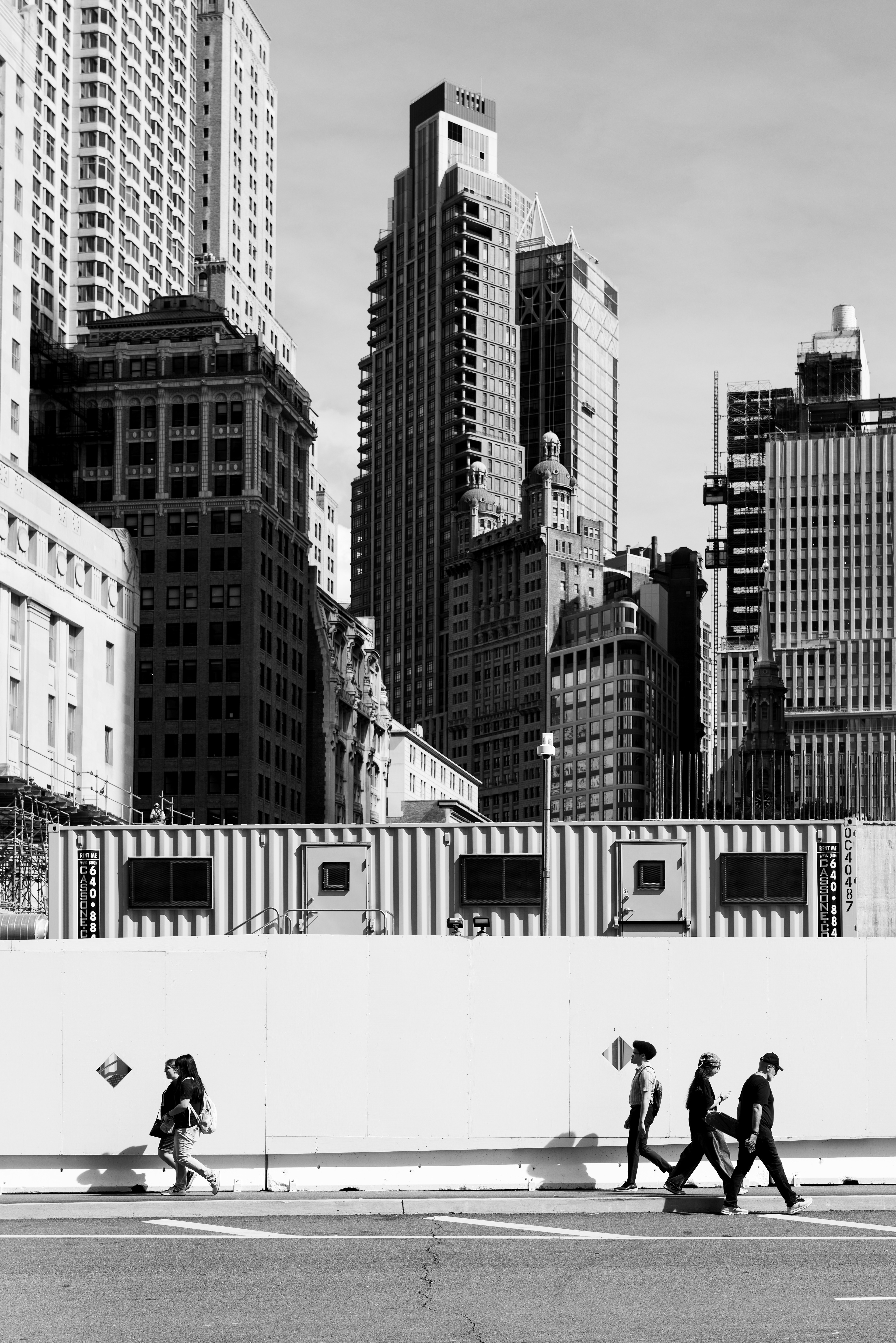 Cette photographie en noir et blanc capture une scène urbaine spectaculaire au cœur de New York City, dans l’environnement architectural dense de Manhattan. L’image met en contraste la monumentalité des gratte-ciel new-yorkais avec la présence quotidienne des piétons dans la rue, offrant une vision à la fois graphique, contemporaine et intemporelle de la ville.  À l’arrière-plan, une succession impressionnante de gratte-ciel et d’immeubles new-yorkais compose un paysage architectural particulièrement dense. De hauts bâtiments contemporains aux façades géométriques côtoient des édifices plus anciens, richement détaillés, révélant plusieurs générations d’architecture de Manhattan. Les lignes verticales des tours attirent immédiatement le regard vers le ciel et donnent à la composition une forte sensation de hauteur et de profondeur.  Au centre de l’image se distingue une imposante tour contemporaine, étroite et élancée, dont la façade sombre contraste avec les bâtiments environnants. À ses côtés apparaissent des constructions historiques aux façades ornées, aux fenêtres répétitives et aux détails architecturaux caractéristiques de l’architecture new-yorkaise. Cette juxtaposition entre architecture historique de Manhattan et gratte-ciel modernes constitue l’un des éléments visuels les plus forts de la photographie.  Au premier plan, une rangée de conteneurs et d’installations de chantier traverse horizontalement l’image. Leur aspect industriel et leurs surfaces métalliques créent une rupture avec les façades monumentales situées derrière eux. La présence du chantier introduit également une notion de transformation permanente : comme souvent à New York, la ville apparaît comme un espace en mouvement, continuellement construit, rénové et réinventé.  Dans la partie inférieure de la photographie, une grande palissade blanche sépare le paysage architectural de la chaussée. Quatre personnes marchent le long du trottoir, créant une échelle humaine face à l’immensité des bâtiments de Manhattan. Les silhouettes des passants sont particulièrement graphiques : leurs corps, leurs vêtements et leurs ombres se détachent nettement sur le fond blanc.  À gauche, une personne avance seule, tandis qu’à droite, plusieurs piétons marchent ensemble dans la même direction. Cette opposition entre une silhouette isolée et un petit groupe de passants apporte du rythme et une véritable narration à la composition.  La lumière est franche et les ombres projetées sur la palissade renforcent l’aspect graphique de l’image. Le choix du noir et blanc élimine les distractions liées aux couleurs et met davantage en valeur les textures, les contrastes, les volumes, les perspectives et les lignes architecturales.  La photographie joue ainsi sur plusieurs niveaux : la monumentalité de New York, l’activité humaine, le chantier, le contraste entre passé et présent et la géométrie des espaces urbains. La route et les lignes horizontales du premier plan s’opposent aux lignes verticales des gratte-ciel, créant une composition équilibrée et très structurée.  L’ensemble évoque une vision cinématographique et documentaire de New York City : une métropole dense, monumentale et en perpétuelle transformation, où les individus semblent minuscules face à l’échelle de l’architecture de Manhattan. New York, New York City, NYC, Manhattan, photographie New York, photo New York, photographie de rue New York, street photography New York, photographie noir et blanc, New York noir et blanc, Manhattan noir et blanc, photographie d'architecture, architecture Manhattan, architecture New York, gratte-ciel New York, skyscrapers New York, New York cityscape, Manhattan cityscape, urban photography, urban landscape, street photography, black and white photography, architectural photography, photographie artistique, photographie contemporaine, paysage urbain, piétons New York, silhouettes, ombres, contraste architectural, ville de New York.  