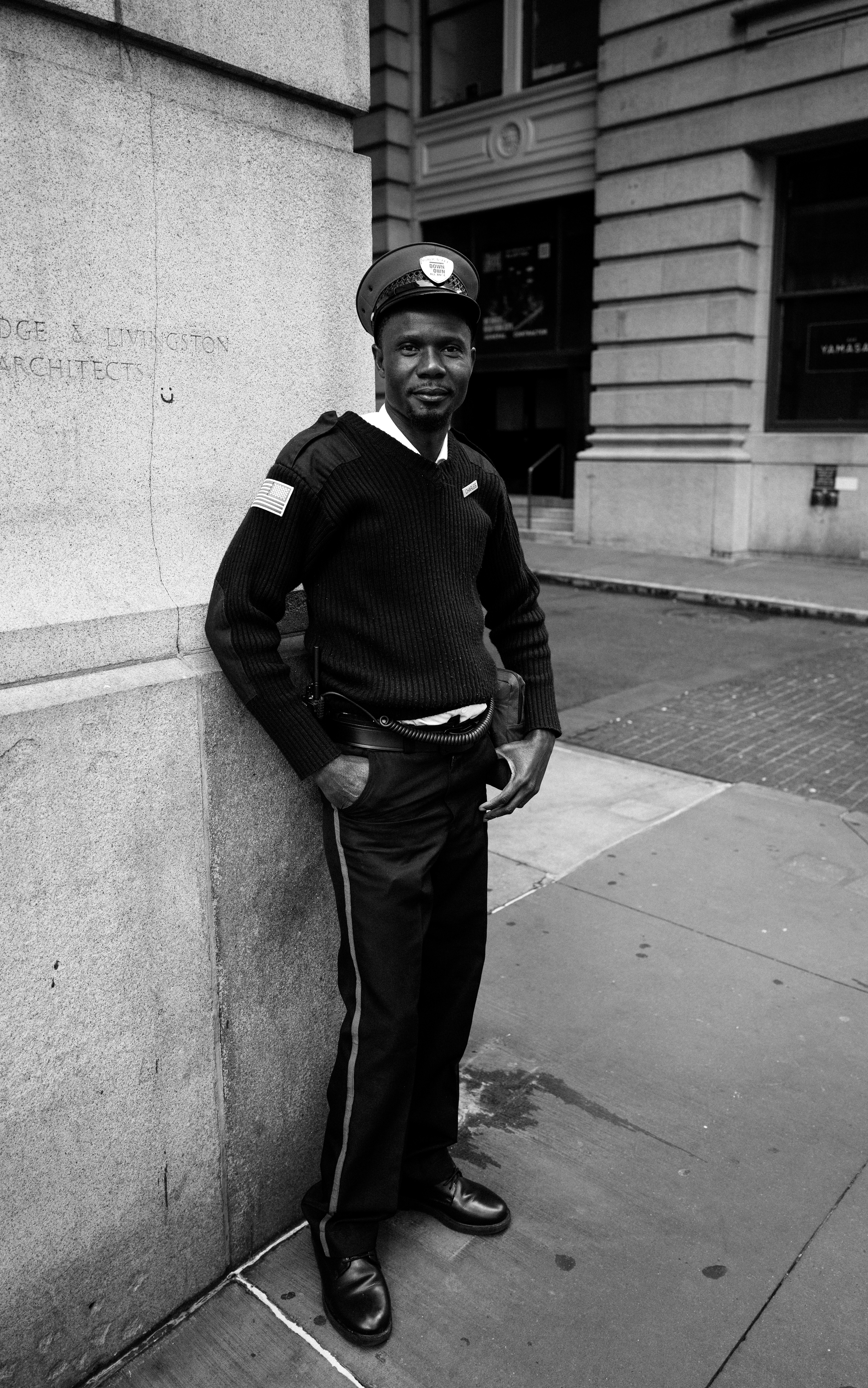 En noir et blanc, ce portrait met en scène un agent de sécurité adossé à l'angle d'un imposant bâtiment en pierre, au cœur d'un environnement urbain. Son attitude est à la fois détendue et assurée : les mains dans les poches, le regard dirigé vers l'objectif, il dégage une présence calme et une confiance naturelle. Son uniforme sombre contraste avec les façades claires qui l'entourent, tandis que les lignes architecturales renforcent la composition et apportent profondeur et équilibre à l'image.  Le choix du noir et blanc met en valeur les textures de la pierre, du tricot de l'uniforme et du bitume, tout en supprimant les distractions de la couleur. La lumière douce révèle les traits du visage avec subtilité, créant une atmosphère intemporelle où se rencontrent le portrait documentaire et la photographie de rue. Cette image célèbre avec sobriété la dignité d'un métier souvent discret, tout en soulignant l'humanité et l'élégance du sujet dans son environnement quotidien. This black-and-white portrait features a security guard leaning against the corner of a monumental stone building in the heart of an urban setting. His relaxed yet confident posture—with his hands in his pockets and his gaze directed toward the camera—conveys both composure and quiet self-assurance. The dark uniform contrasts beautifully with the light-colored architecture, while the strong architectural lines add depth, balance, and structure to the composition.  The monochrome treatment emphasizes the rich textures of the stone, the knit fabric of the uniform, and the pavement, removing the distraction of color and allowing the viewer to focus on expression and form. Soft natural light gently reveals the subject's facial features, creating a timeless atmosphere that blends documentary portraiture with street photography. The image pays tribute to the dignity of an often-overlooked profession while highlighting the humanity and quiet elegance of its subject within his everyday environment.