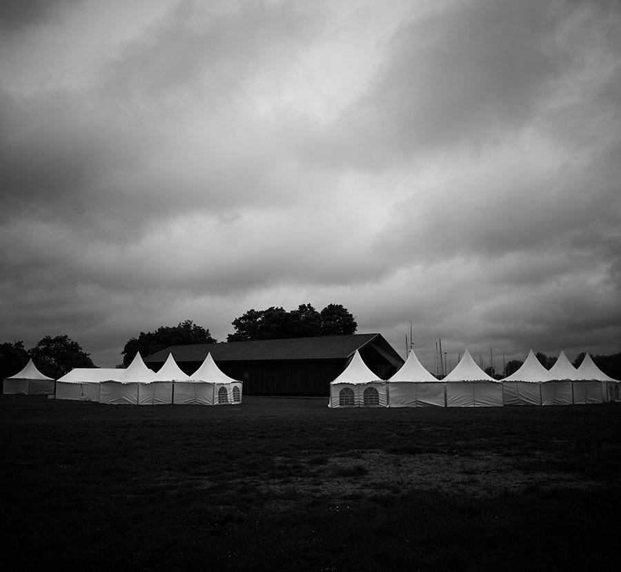 Sous un ciel chargé où les nuages semblent suspendre le temps, une série de tentes blanches se détache avec force dans un paysage presque monochrome. Leur géométrie régulière contraste avec les formes organiques des arbres et l'immensité du ciel, créant un équilibre entre ordre humain et nature. La lumière diffuse, filtrée par une épaisse couverture nuageuse, accentue les textures et renforce l'atmosphère silencieuse qui imprègne la scène.  Le choix du noir et blanc met en valeur les contrastes, les volumes et les nuances du ciel, tout en invitant le regard à s'attarder sur la simplicité des formes. Cette photographie explore la rencontre entre architecture éphémère et paysage, où l'attente, le calme et une légère tension météorologique deviennent les véritables sujets de l'image. Beneath a heavy sky where the clouds seem to suspend time, a series of white tents stands out against an almost monochromatic landscape. Their clean, repetitive geometry contrasts with the organic shapes of the trees and the vastness of the sky, creating a balance between human order and nature. The soft, diffused light filtered through the thick cloud cover enhances textures and reinforces the quiet atmosphere that permeates the scene.  The choice of black and white emphasizes contrast, form, and the subtle tonal variations of the sky, inviting the viewer to linger on the simplicity of the composition. This photograph explores the meeting point between temporary architecture and the landscape, where anticipation, stillness, and the quiet tension of approaching weather become the true subjects of the image.