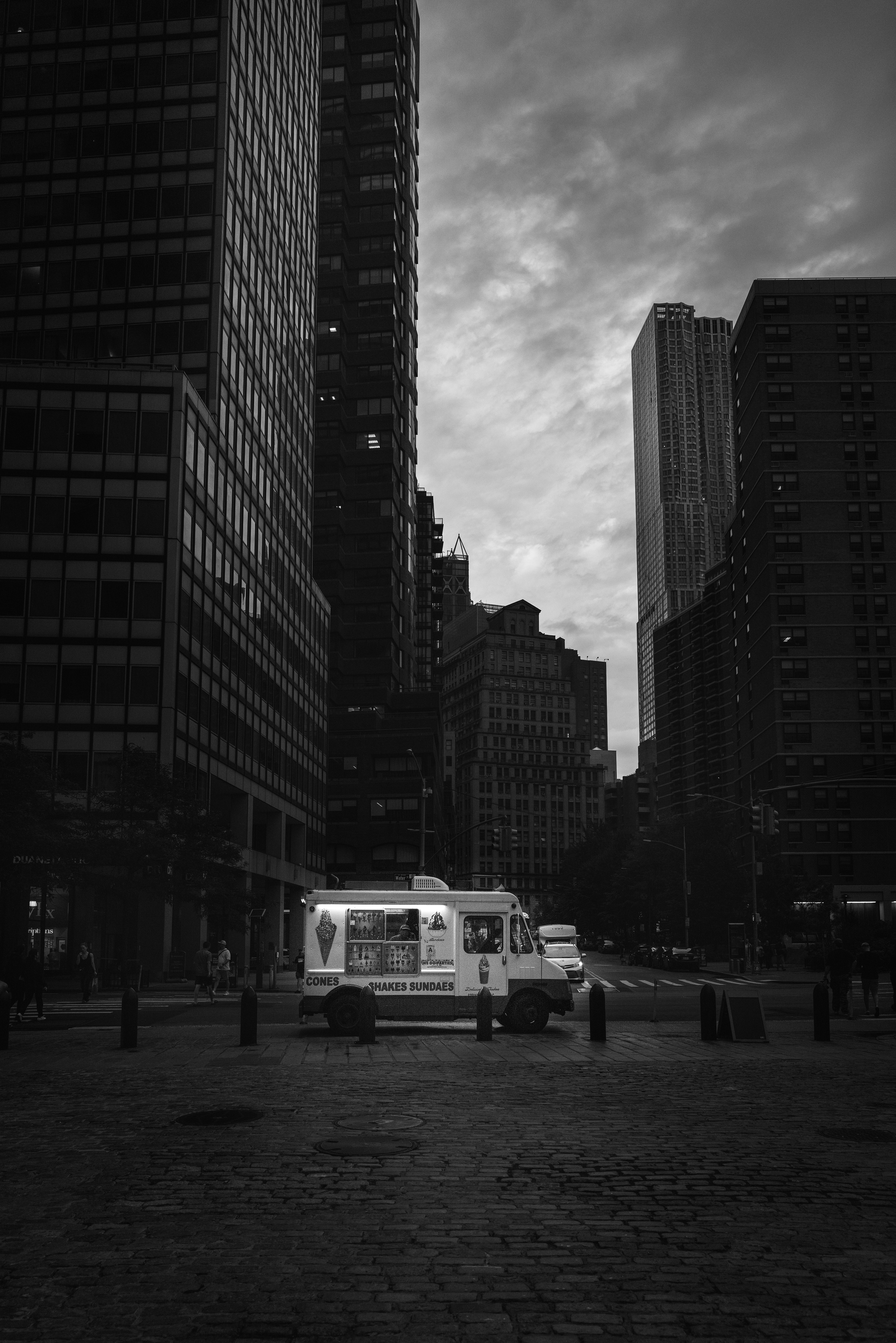 Cette photo en noir et blanc, réalisée avec un Leica Q2 dans les rues de Manhattan, montre un camion blanc qui contraste avec les ombres de la ville, au milieu des immeubles. This black-and-white photograph, taken with a Leica Q2 in the streets of Manhattan, features a white truck standing out against the city's shadows, surrounded by towering buildings.