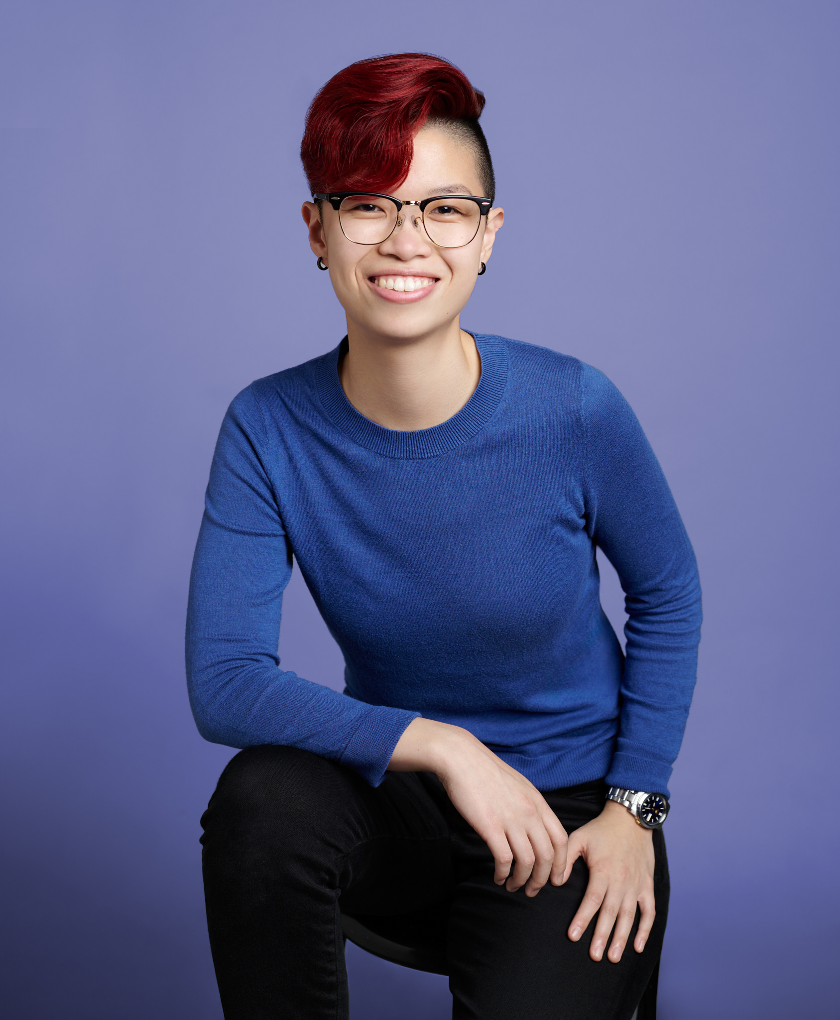 A portrait of Zenna Wong, a smiling young asian woman with short vibrant red hair wearing a rich blue long sleeve shirt and black jeans. She sits on a stool with one knee up against a plain purple background.