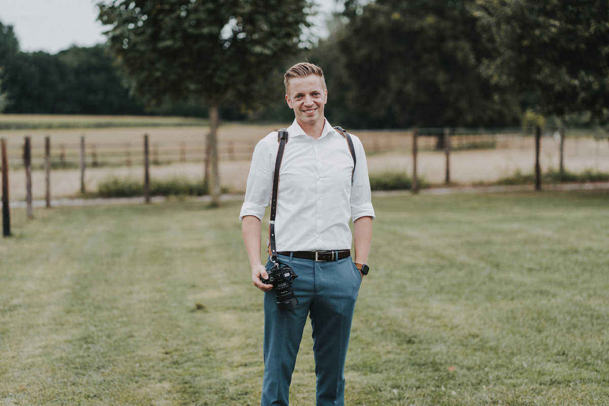 Alt-Text: Ein Hochzeitsfotograf steht lächelnd auf einer grünen Wiese vor einem unscharfen Hintergrund mit Bäumen und einem Zaun. Er trägt ein weißes Hemd mit hochgekrempelten Ärmeln und eine blaue Hose. Um seine Schultern ist ein Kameragurt befestigt, und in seiner rechten Hand hält er eine Kamera.