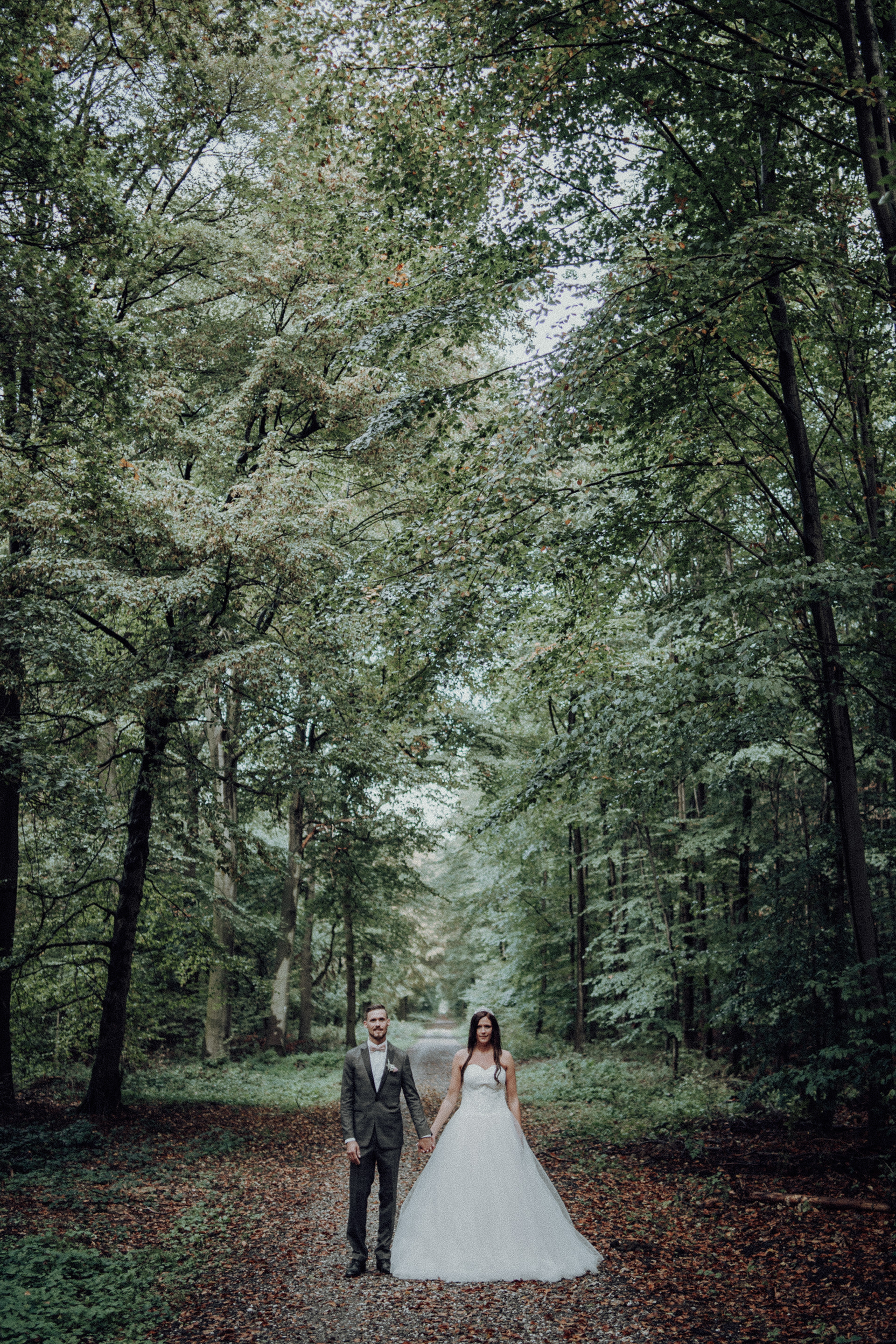 Brautpaar steht straight zu Kamera auf Waldweg.