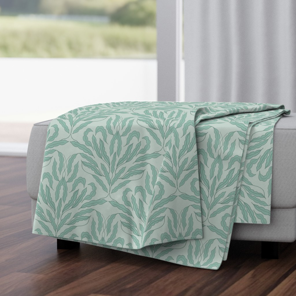 A folded throw blanket draped over a light grey sofa, featuring a soft mint‑teal botanical pattern. Warm daylight filters through a nearby window, highlighting the fabric’s gentle texture in a calm, cozy home setting.