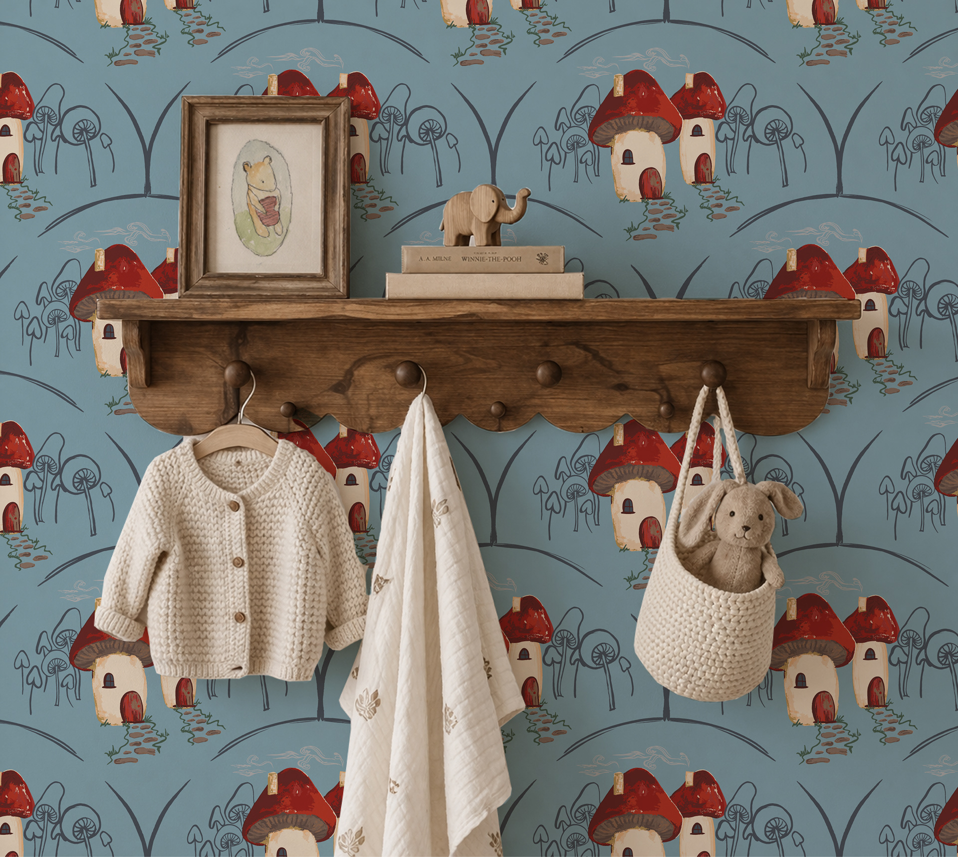 Nursery wall shelf against blue mushroom‑themed wallpaper featuring illustrated mushroom houses and trees. The shelf holds a framed bear illustration, stacked Winnie‑the‑Pooh books, and a small elephant figurine. Below hang a knitted baby cardigan, patterned cloth, and a crocheted basket with a stuffed bunny, creating a cozy, storybook atmosphere.
