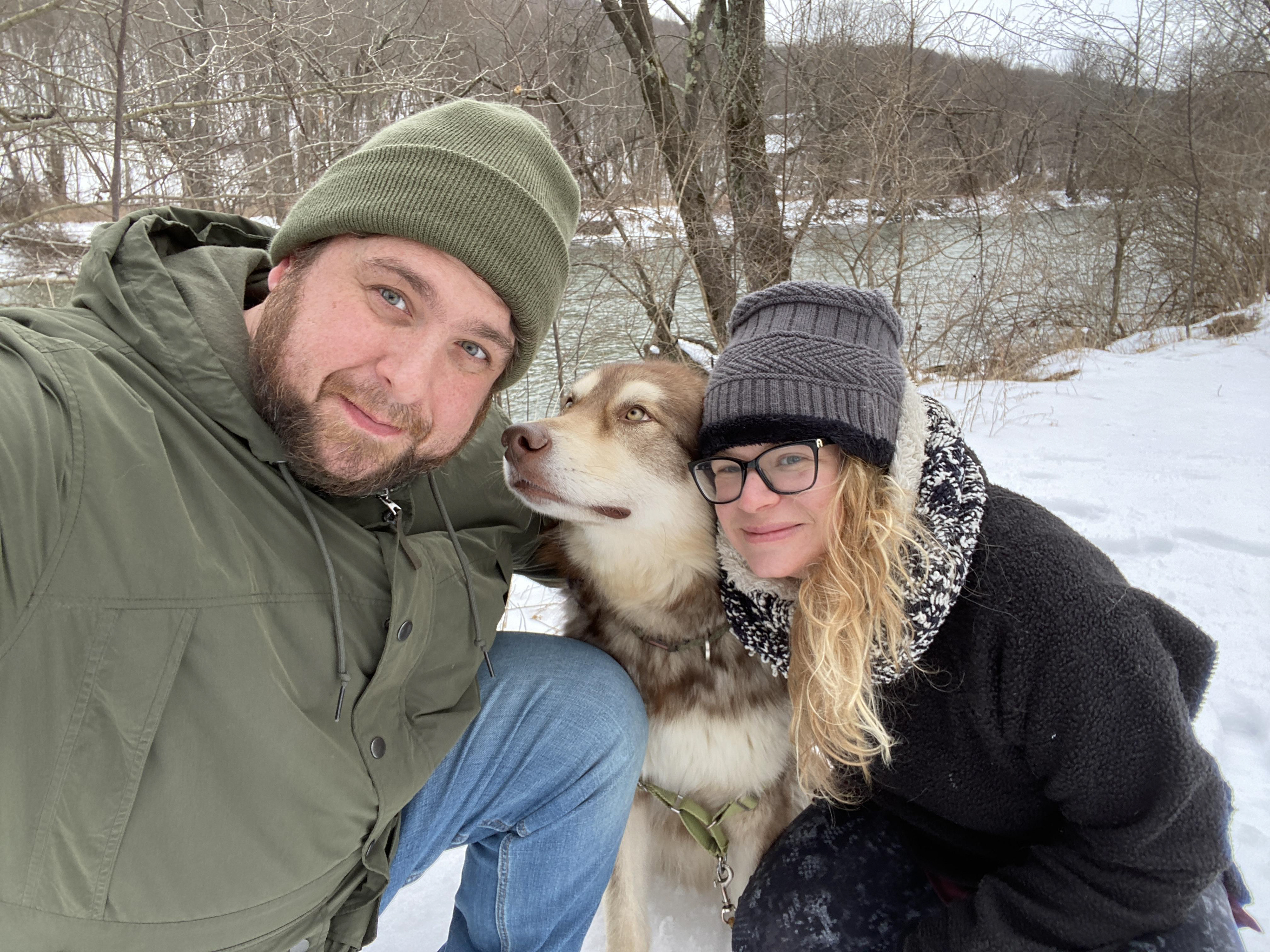 Christopher Starr smiling alongside his wife and their Malamute dog against a snowy backdrop, showcasing a warm family moment outdoors.