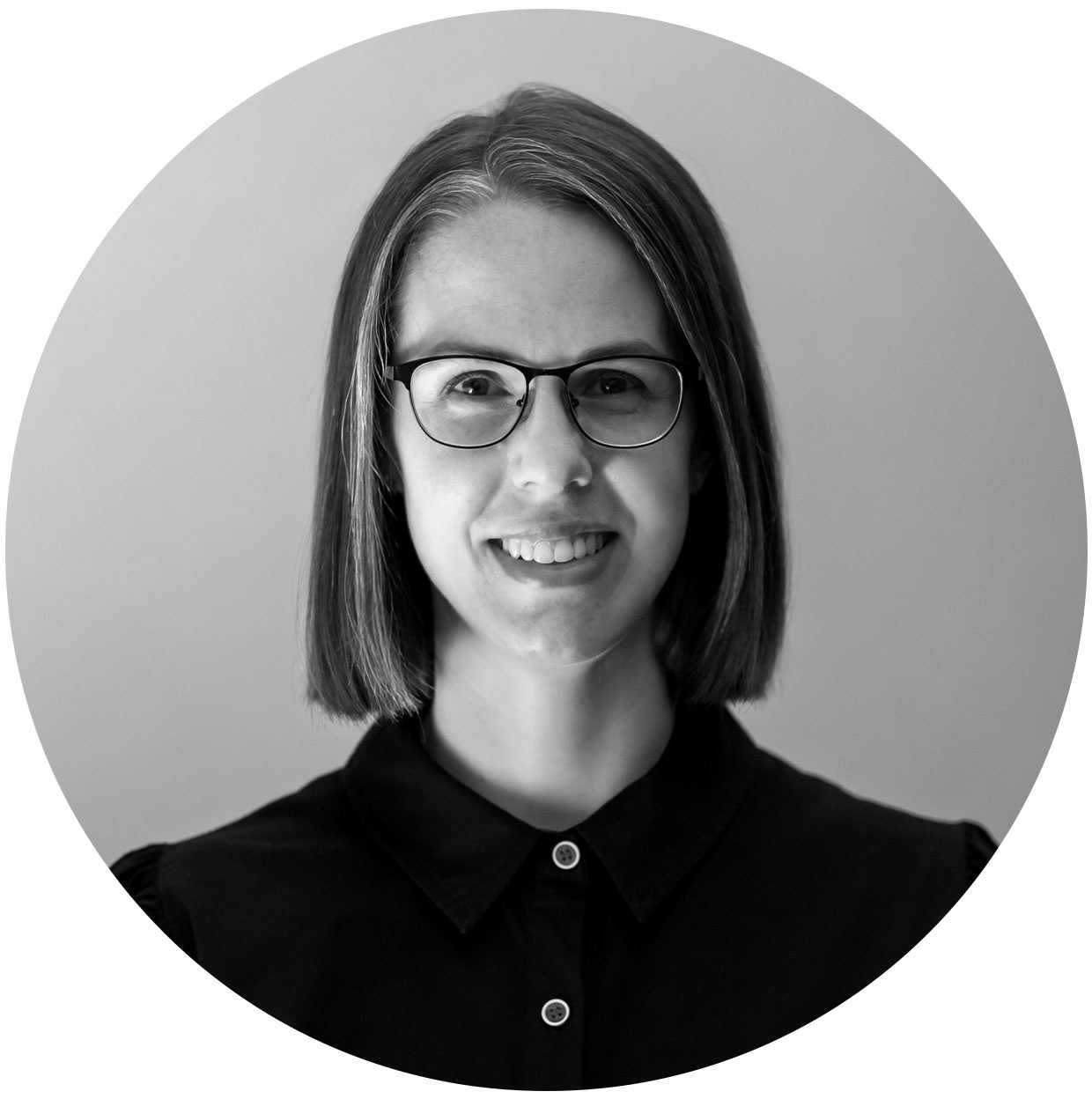 A black and white portrait of Wilna Combrinck. She is smiling, wearing a dark button-up dress with a collar. She has shoulder-length hair and wears glasses.