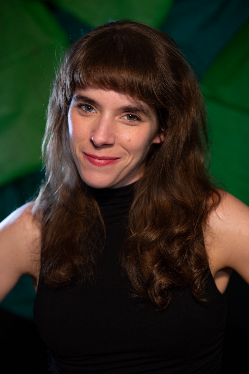 Portrait of Jessye from the waist up, smiling at the viewer. She has brown hair that just passes her shoulders with bangs and is wearing a black tank top.