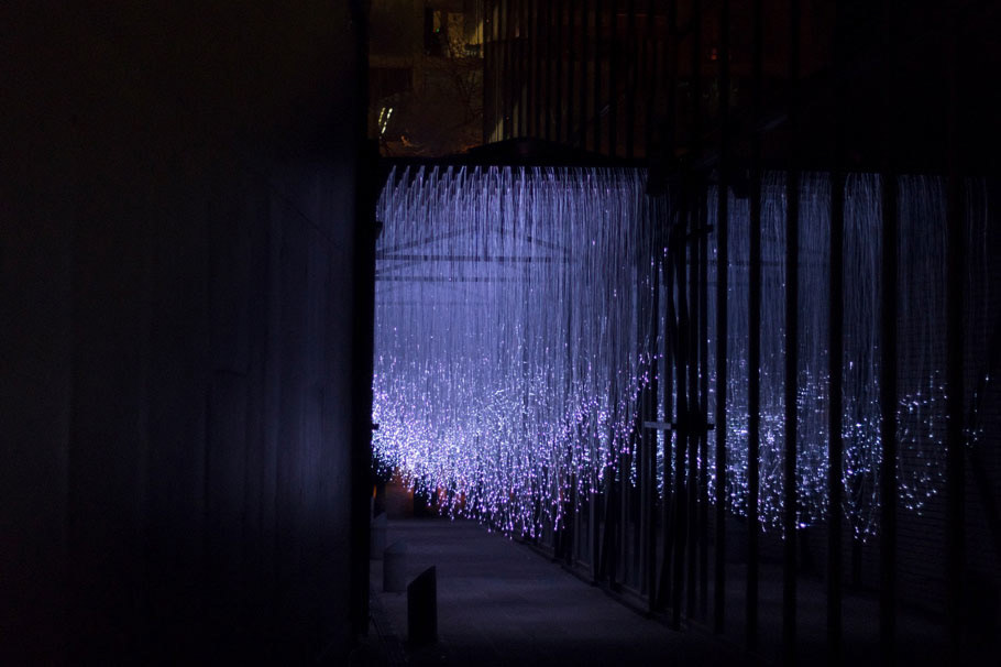 A full view of the fiber optic light installation, held together by a black wooden structure placed in a walkway between a wall on the left and a glass facade on the right.
