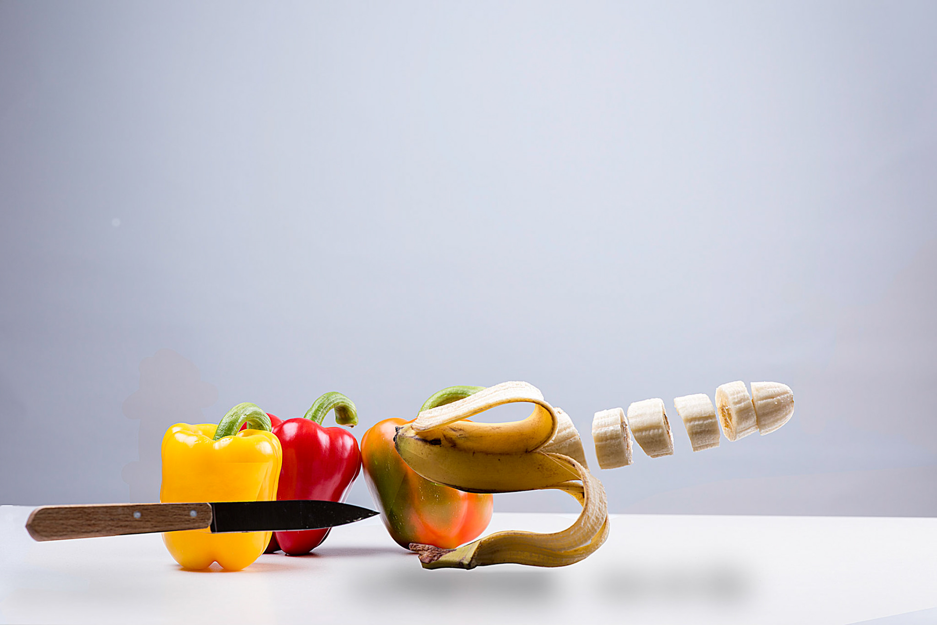 Banana on the run for a kitchen knife in front of a red yellow and green paprika while already been half peeled and sliced