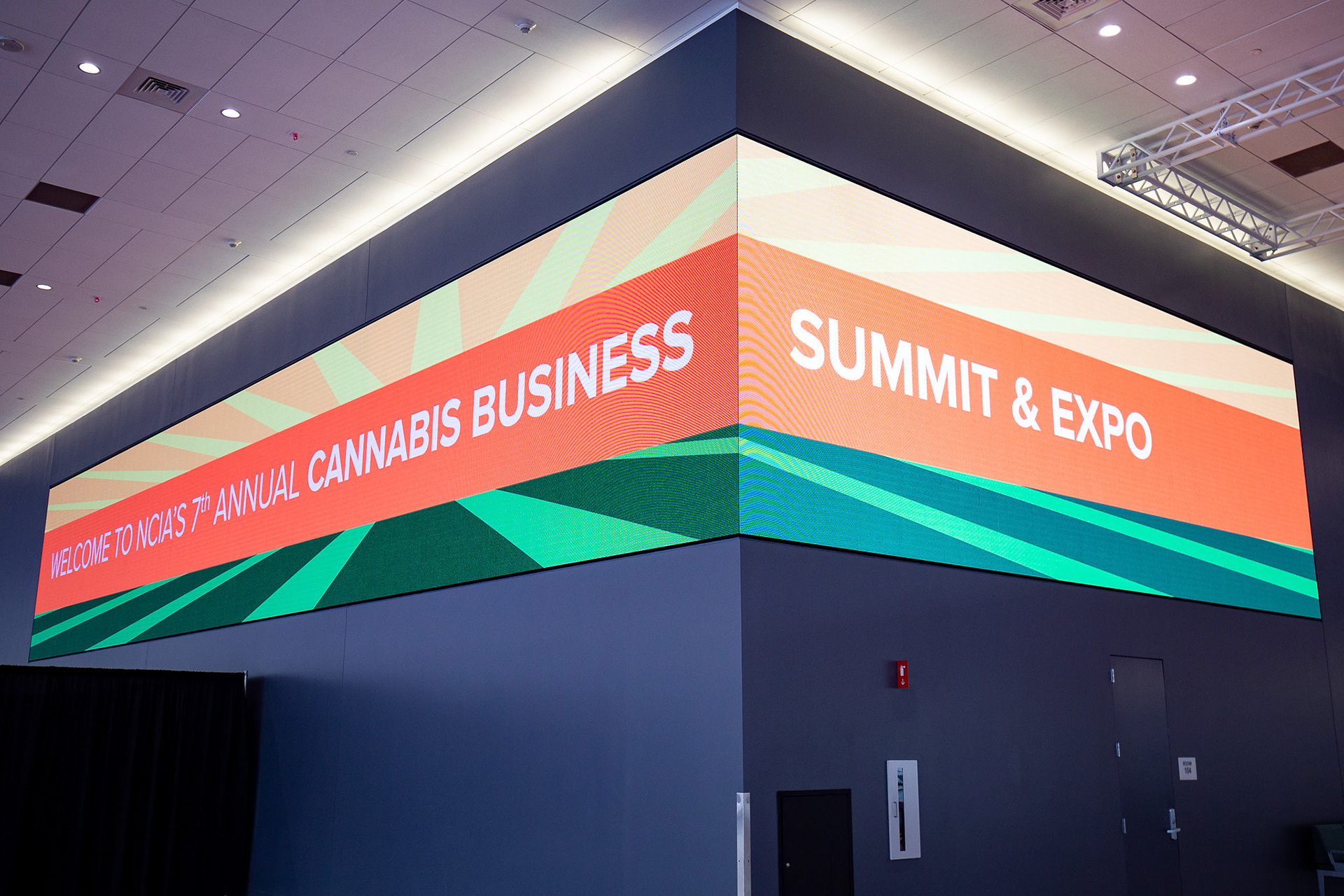 Attendees speaking with NCIA staff at an illuminated engagement counter with green and orange graphics and a branded backdrop at the 2021 Cannabis Business Summit & Expo.