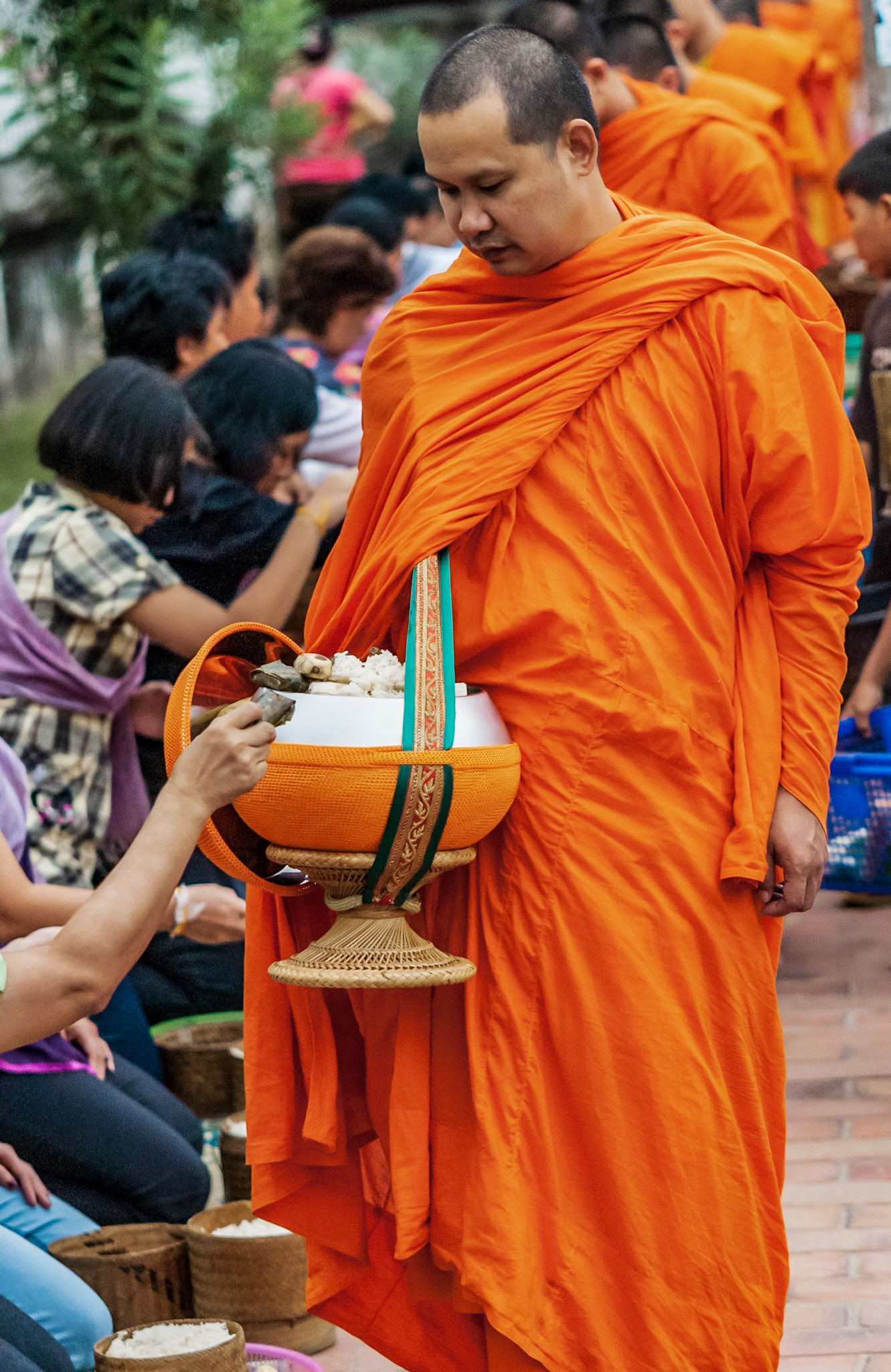 Giving alms to monks fosters a spiritual connection for Buddhists. The seated participants add their gifts to the monks’ bowls, connecting with the spiritual realm and showing humbleness and respect while earning merit on the pathway to enlightenment.