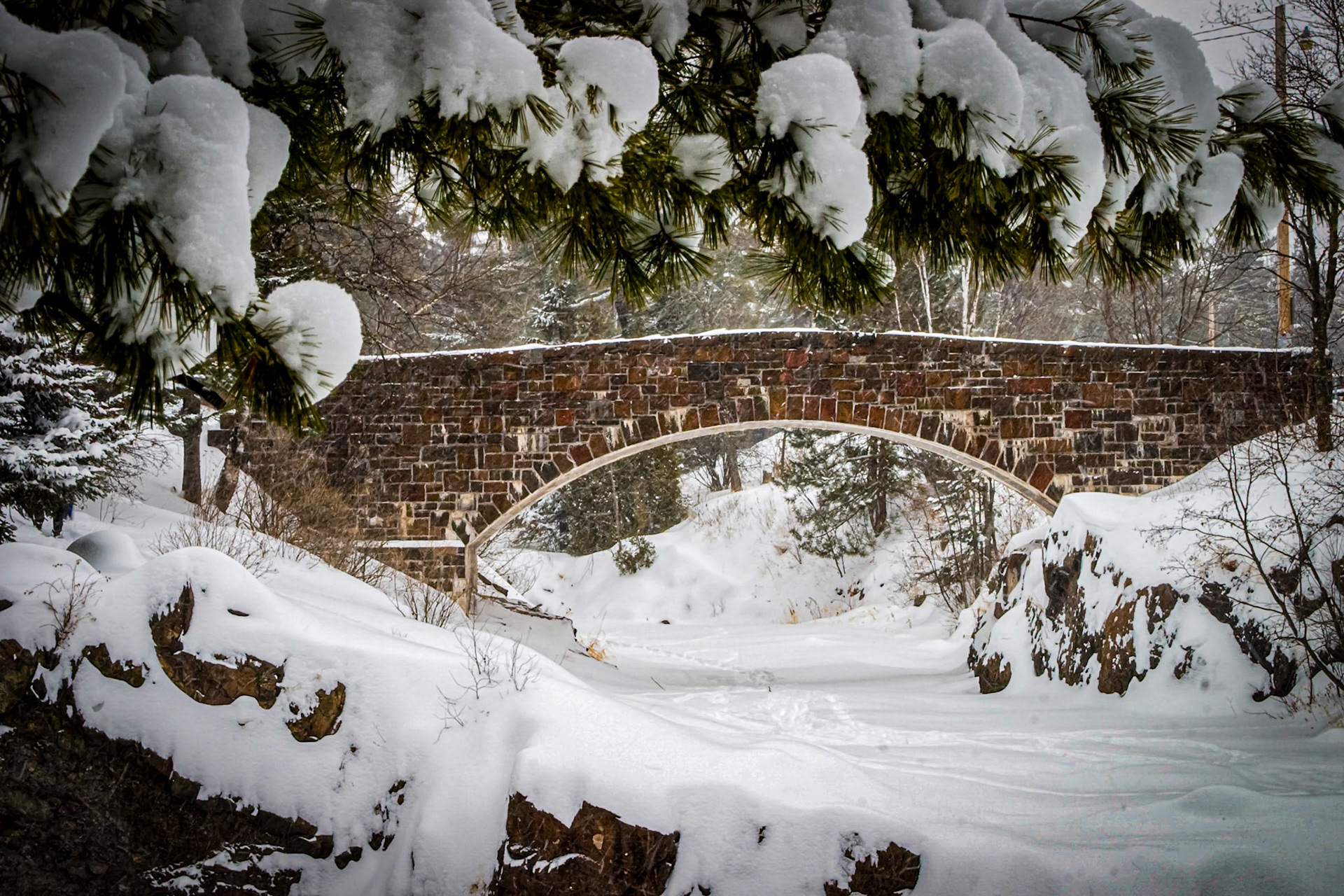 January 26 - Winter PassageA blanket of freshly fallen snow covers the landscape around the Amity Creek Stone Arch Bridge in Duluth. This small pedestrian bridge is a work of art built in the 1930's by Depression-era stone masons. Hand-laid from local stone and designed to blend into the gorge, it is a tribute to a long-lost craft. The heavy snow cover muffles every sound, yet you can still hear the hidden water flow beneath the ice. The joy of winter is the quiet peacefulness surrounding you on a walk up a frozen Lester River. The occasional chirp of a chickadee or squawk of a raven breaks the silence, only to return to peaceful quiet. You see this scene, and you can't help but believe that it was crafted by the hand of God just for this moment to interrupt your day.You keep him in perfect peace, whose mind stays on you because he trusts you. Isaiah 26:3The only peace and safety in this world is hope and faith in our Lord and Savior, Jesus Christ. Walk in the truth and enjoy today.