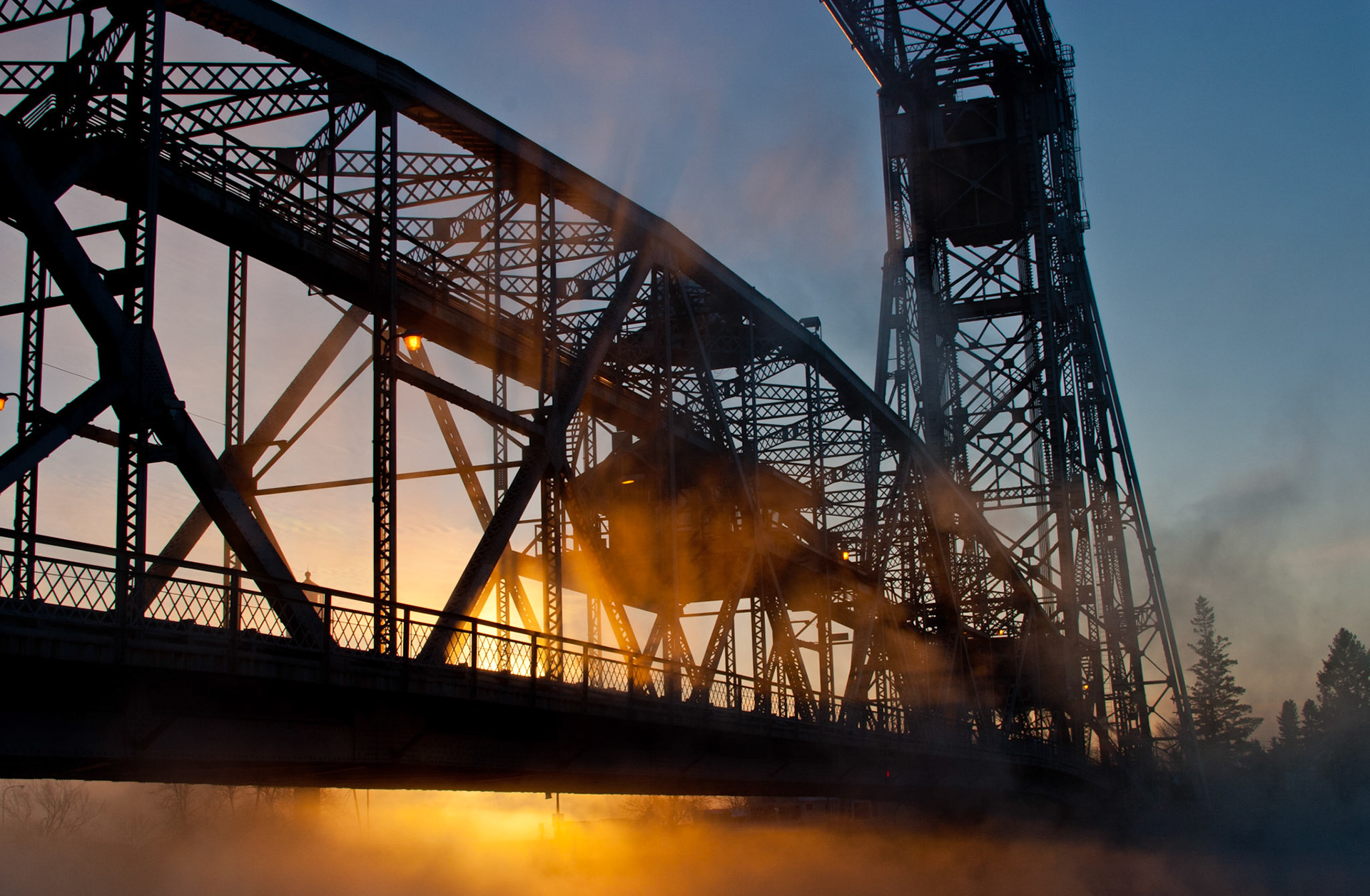 December 22 - Cold Iron, Warm SunriseSea Smoke rises through the iron gridwork of the Duluth Aerial Lift Bridge on this -20F December morning.From the rising of the sun to its setting, the name of the Lord is to be praised! Psalm 113:3
