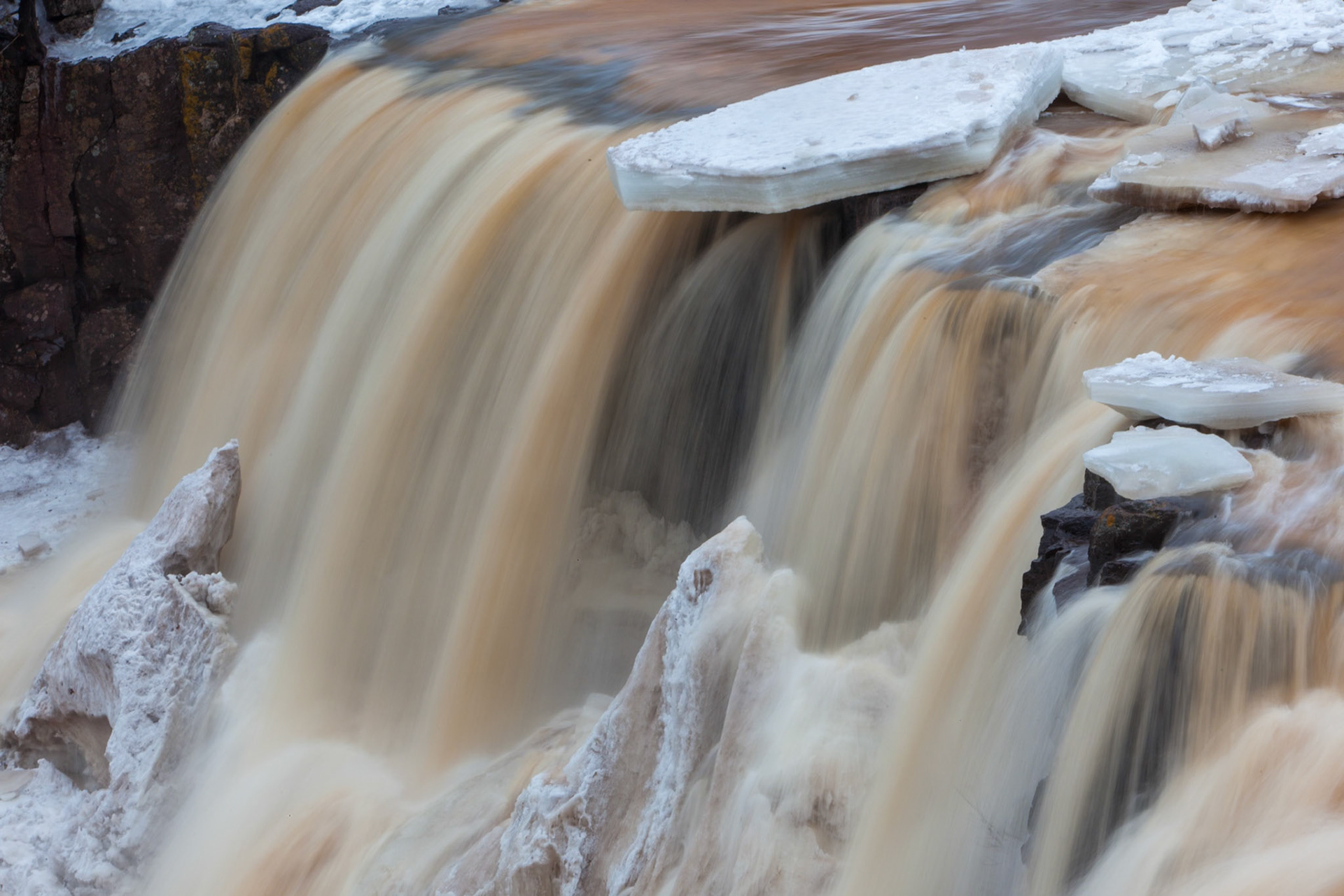 March 25 — Spring BreakupGooseberry Falls State Park is one of the finest places to witness spring’s arrival. Thick shelves of winter ice cling to the edges of the falls, suspended above the rushing Gooseberry River as they await their return to Lake Superior just downstream.On the North Shore, spring begins with a whisper — longer light, subtle warmth — and then suddenly it roars. Ice fractures and crashes against dark basalt below. Snowmelt swells the river, tinting it with the color of the land. The air fills with mist and thunder, a scene impossible to ignore.Power and beauty combine in this season of release. What seemed frozen and immovable only days before gives way to unstoppable flow. Spring can no longer be restrained.“For by him all things were created, in heaven and on earth, visible and invisible… all things were created through him and for him.” — Colossians 1:16Whether you bring a camera or stand still, Gooseberry in spring engages all your senses. The thunder, the spray, the scent of thawing earth — it humbles the spirit and refreshes the soul. Creation is always at work, even when we think it stands still.