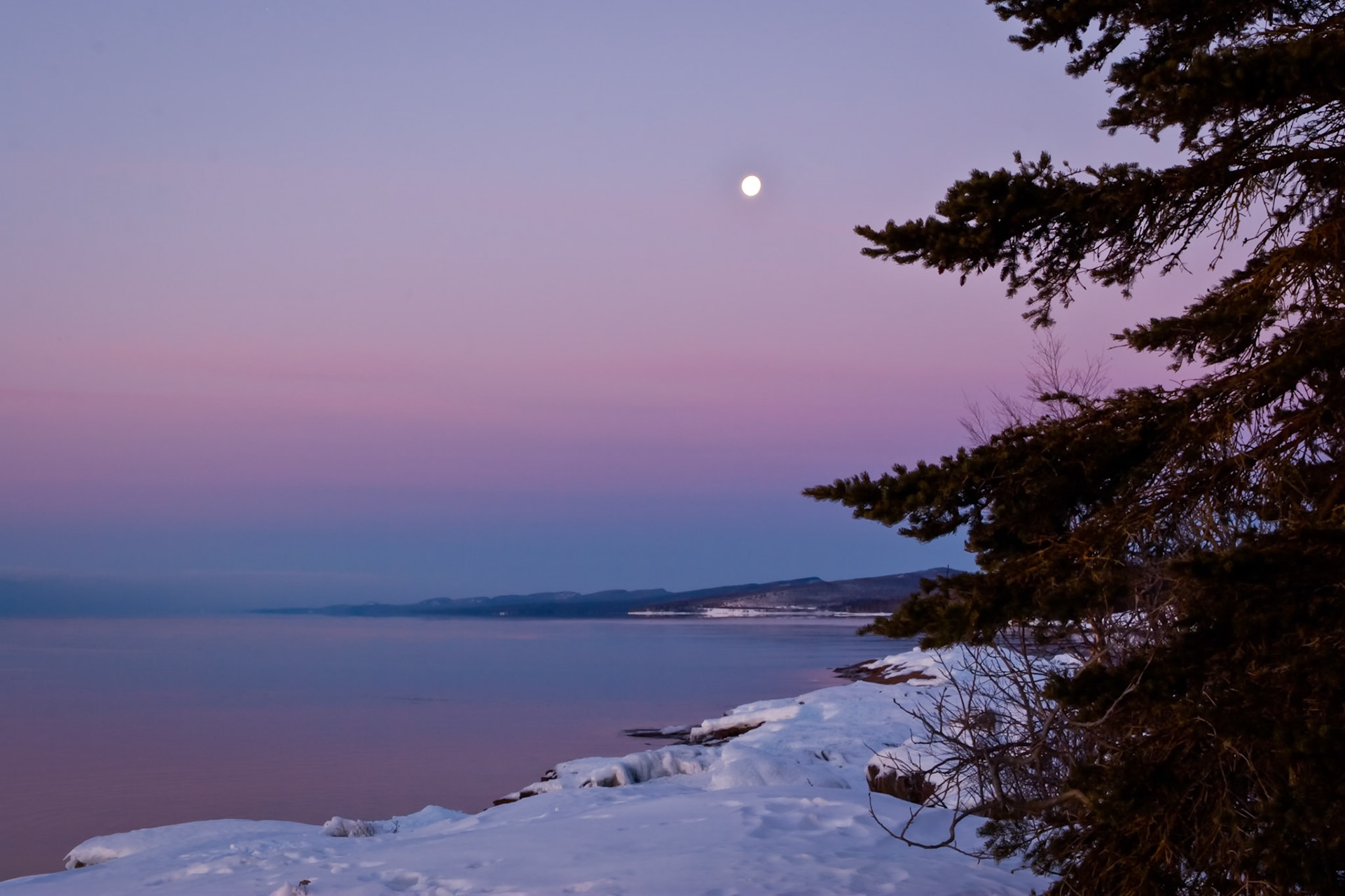 February 24th - Magenta ShoresThe setting moon slowly descends over the snow-covered shores of Lake Superior near Grand Marais. Trees, rocks, snow, and soft winter light come together to form a picturesque landscape during this brief and peaceful moment of the day.It is a good feeling to see open water after a long winter, and it is refreshing to realize that daylight and nighttime hours will soon stand in balance once again.The moon drops toward the horizon as the Earth continues its daily journey, unnoticed as time marches on. Watching the vastness of the scene and the slow motion of the moment helps put things into perspective — reminding us that we are not the center of the universe, but passengers on a small planet created by a great God.Like cold water to a thirsty soul, so is good news from a far country. Proverbs 25:25Hope comes in many forms, and a quiet February landscape along the shores of Lake Superior offers a perfect beginning in the hands of our loving God.