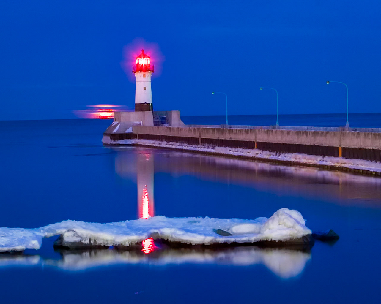February 12 - ReflectionsA February full moon lifts above the horizon on a calm winter evening. The North Pier Lighthouse — standing watch for more than 115 years — reflects softly across glassy water, joining moonlight and shore ice in layers of deep winter blue.Cold, calm water acts like natural glass. With fewer surface ripples and greater density, winter conditions create reflections far sharper than those seen during warmer months, turning the harbor into a mirror of light and shadow.Since the Winter Solstice on December 21, the Northland has gained 78 minutes of daylight. Each passing day lifts the sun a little higher, strengthening its warmth and revealing new textures in ice, water, and sky — gentle signs that winter’s long hold is slowly loosening.“And we all, who with unveiled faces contemplate the Lord’s glory, are being transformed into his image with ever-increasing glory.” — 2 Corinthians 3:18We have the ability to reflect — through memories, photographs, journals, and stories. Some reflections are good, others are more difficult. The ability to forget can also be a gift. Today is a fresh, new day that will soon become part of the past — make it one worth remembering.