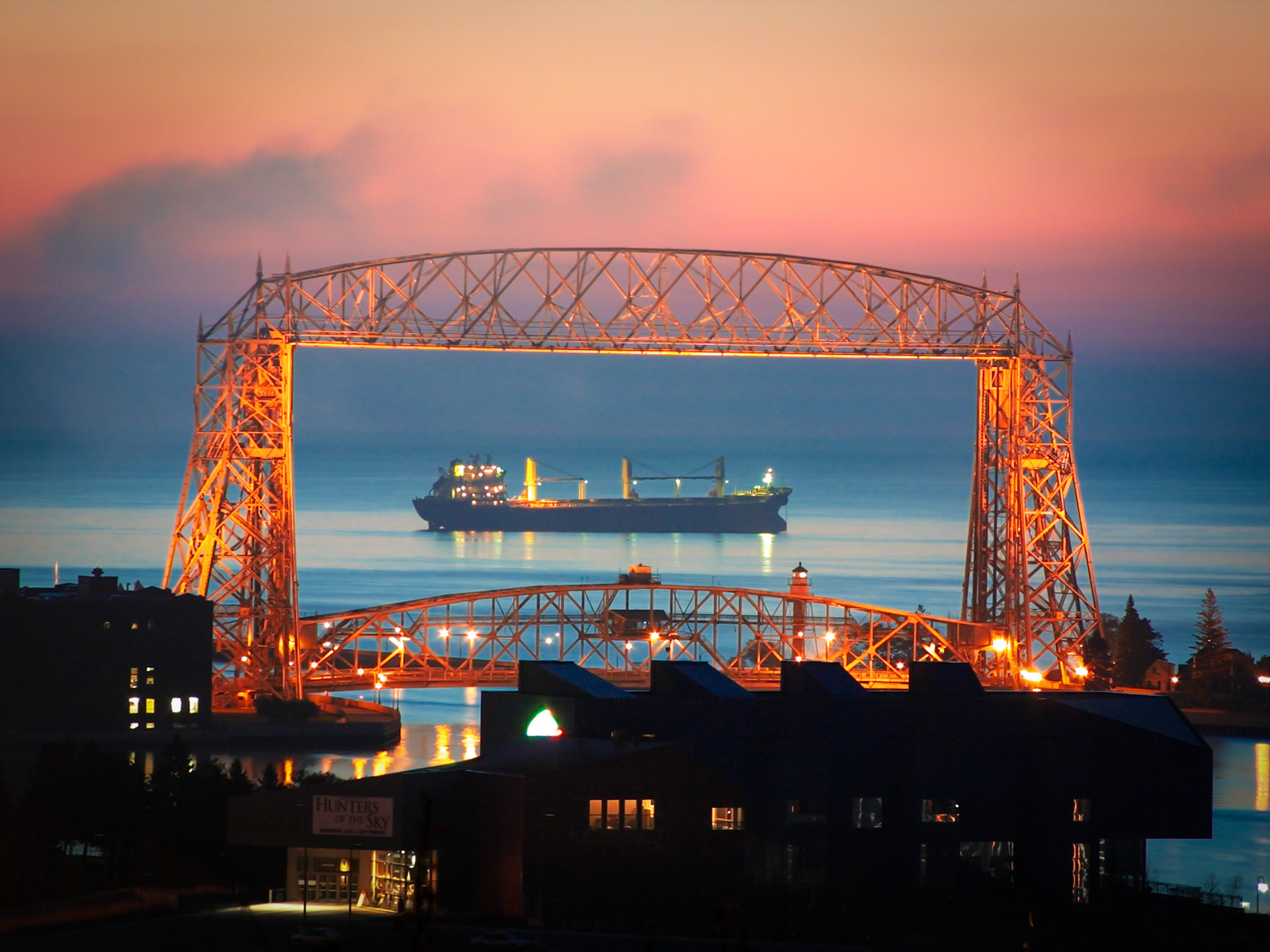 November 16 - Pastel Morning A freighter waits at anchor for its turn to load grain in Duluth.Framed by the Aerial Bridge, the large boat has purposely moved into position for a perfect photo opportunity.The still waters of Lake Superior reflect the sky's transformation, creating a breathtaking scene. It's a moment of tranquility before the hustle and bustle of the day begins.For with you is the fountain of life; in your light do we see light. Psalm 36:9