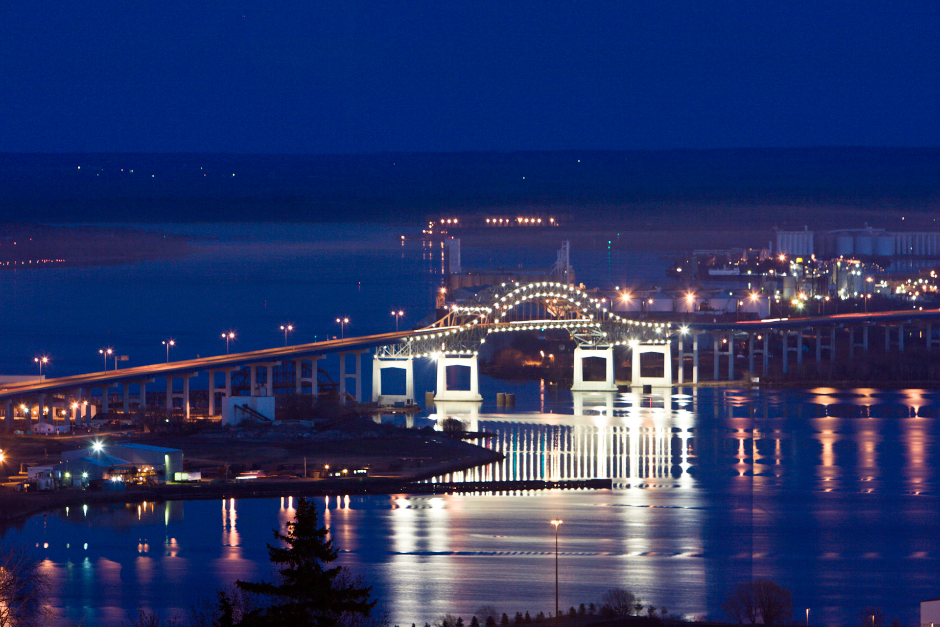 April 21st: Blatnik Bridge Night Light - Lights from the Blatnik Bridge reflect off the St. Louis Bay just after twilight, and a peaceful calm settles across the bay.The Blatnik Bridge is a truss arch bridge spanning Saint Louis Bay on Interstate Highway 535, connecting Duluth, Minnesota, and Superior, Wisconsin. It is named in honor of Congressman John A. Blatnik and opened to traffic in 1961. The bridge is 7,980 feet long and 120 feet above the water. The bridge also provides a stunning view of Saint Louis Bay, especially during sunrise and sunset."Trust in the Lord with all your heart and lean not on your own understanding; in all your ways submit to him, and he will make your paths straight." - Proverbs 3:5-6This scripture reminds us to trust God's guidance and not rely solely on our understanding. Just like a bridge can help us overcome a physical obstacle, trusting in God can help us navigate life's challenges and lead us down the right path.