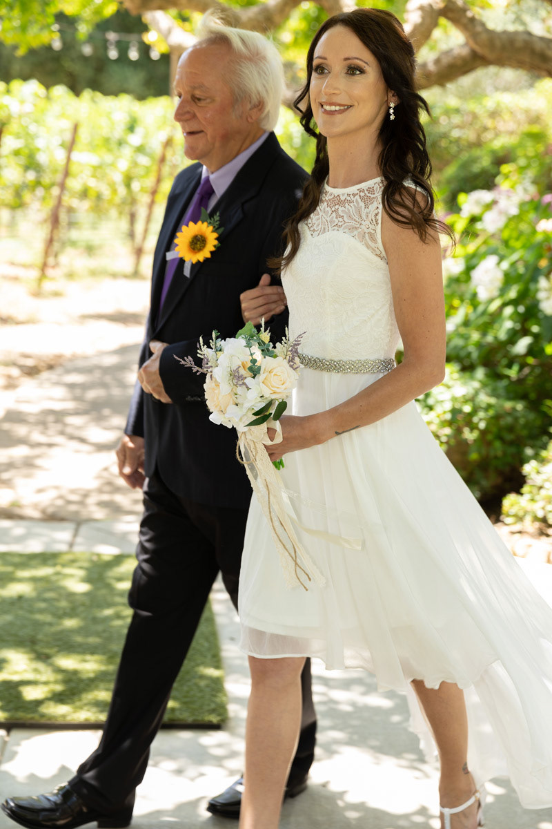 father-with-sunflower-boutonniere-and-bride-with-white-rose-bouquet-stride-arm-in-arm-in-napa-private-vineyard