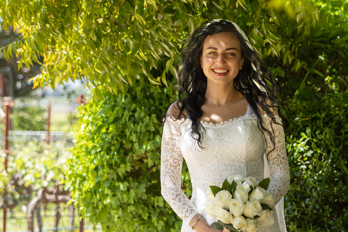 bride-with-long-black-wavy-hair-in-white-lace-dress-holds-a-bouquet-of-white-roses-in-front-of-a-vineyard