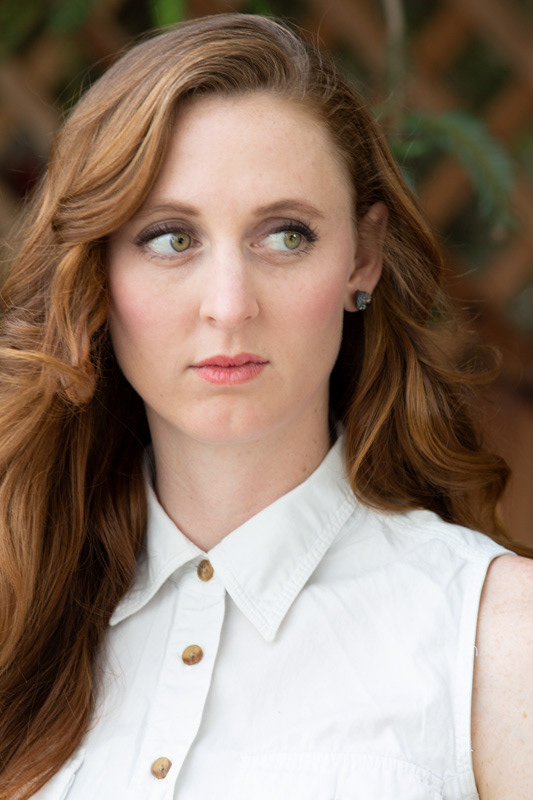 young-woman-with-green-eyes-and-long-wavy-red-hair-wears-a-white-button-up-sleeveless-top-her-eyes-look-off-to-the-side