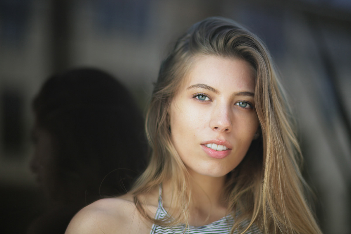Young woman-with-highlighted-blond-hair-and-blue-eyes-wears-a-white-and-blue-striped-halter-top-and-leans-against-a-dark-window