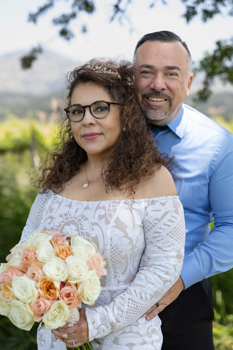 bride-with-long-curly-brown-hair-wers-a-lace-gown-and-carries-a-white-andorange-rose-bouquet-her-back-against-the-grooms-chest-with napa-valley-view-in-background