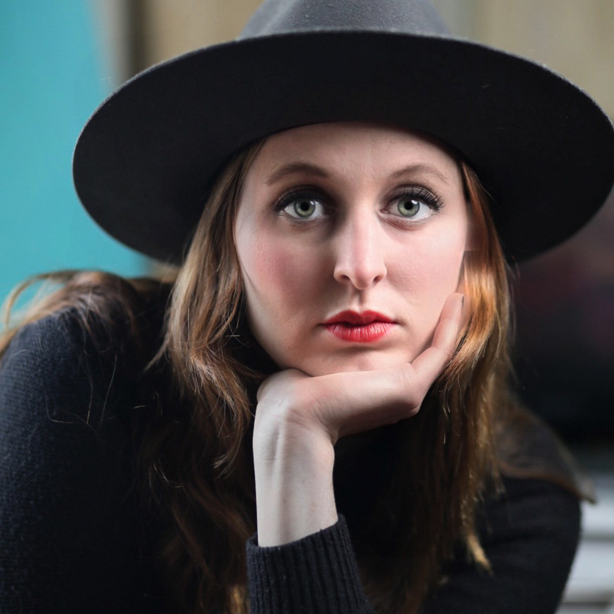 woman-with-deep-red-lips-and-green-eyes-peers-out-from-under-a-black-wide-brim-hat-with-chin-resting-in-palm 