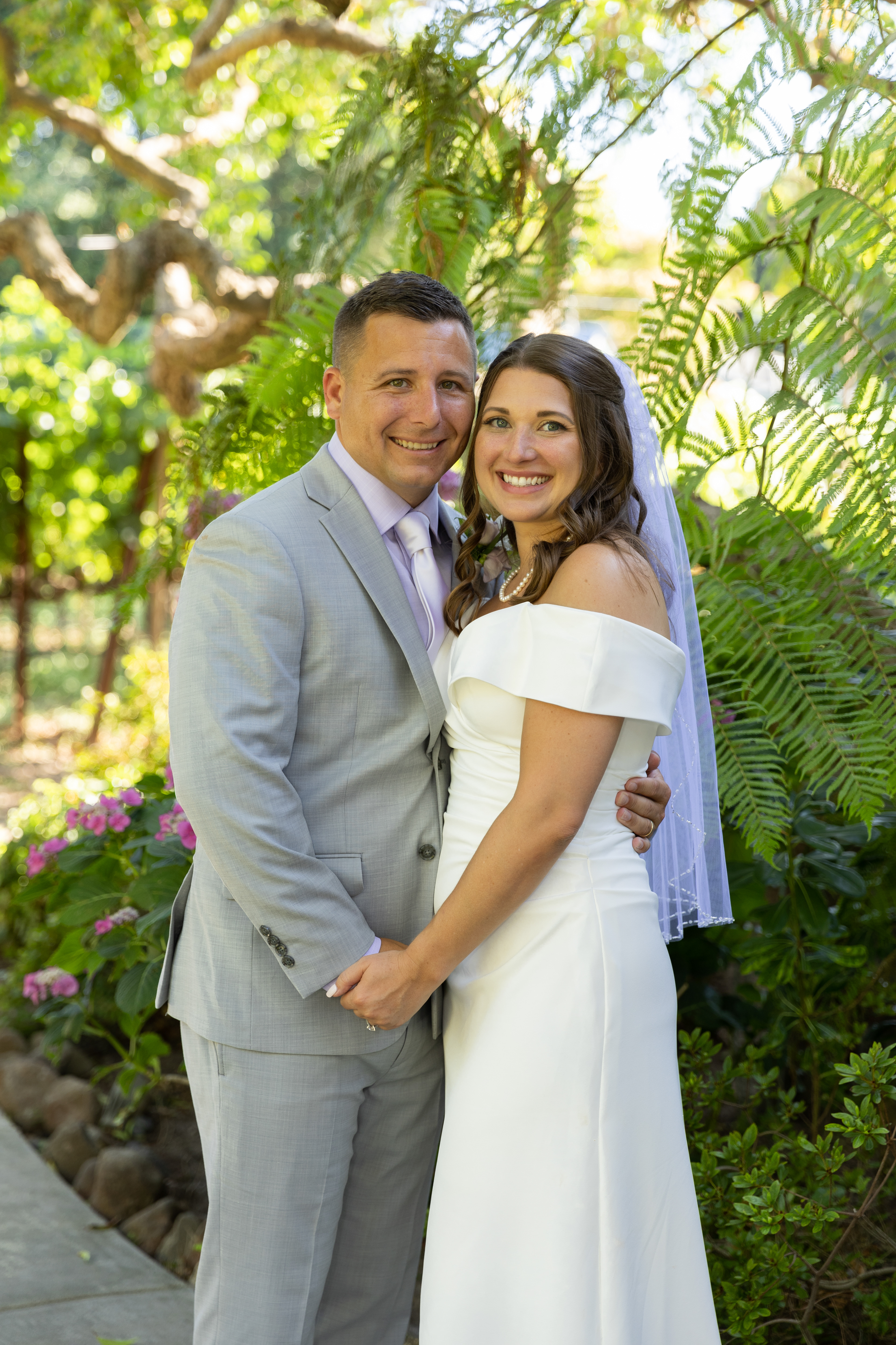 Groom-in-grey-suit-and-lavendar-shirt-holds-the-hand-the-bride-centered-lush-green-foliage-background