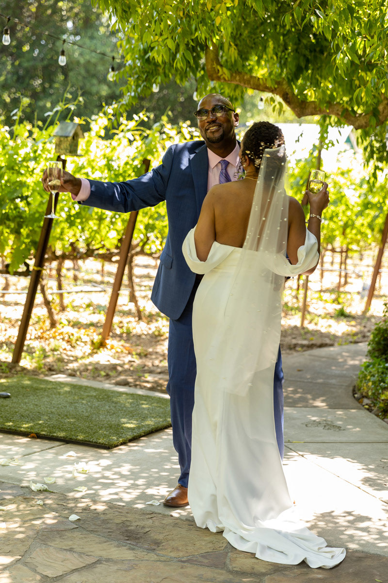 a-groom-wears-a-blue-suit-holds-a-glass-of-champagne-while-dancing-with-the-bride-in-a-wine-country-garden