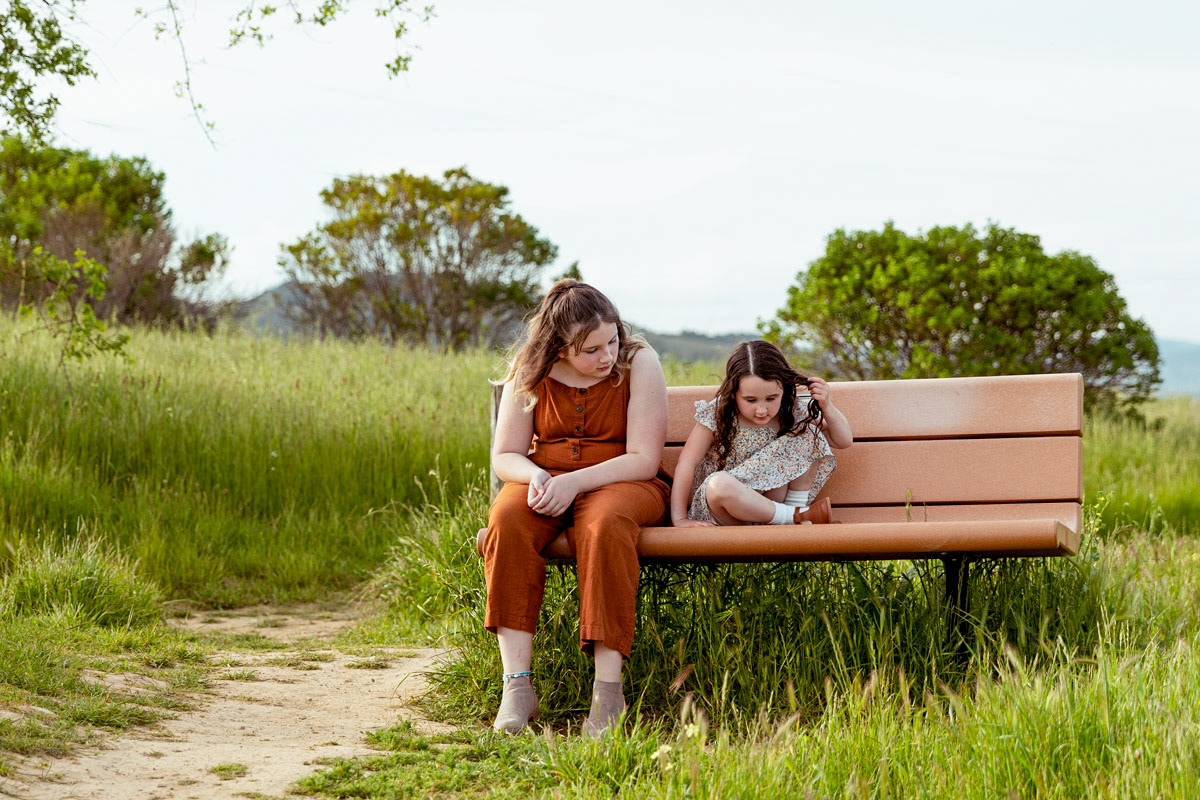Two-girls-onesmall-one-tall-sit-on-a-Napa-park-bench-watching-something-in-the-tall-grass-in-front-of-them