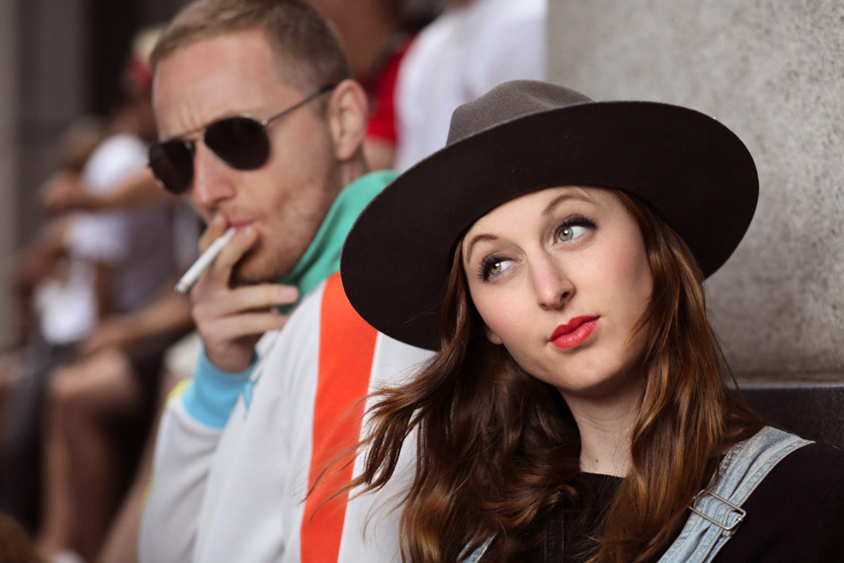 woman-with-big-green-eyes-wears-long-red-hair-red-lipstick-and-a-wide-brim-black-hat-with-man-in-sunglasses-smoking-over-her-shoulder