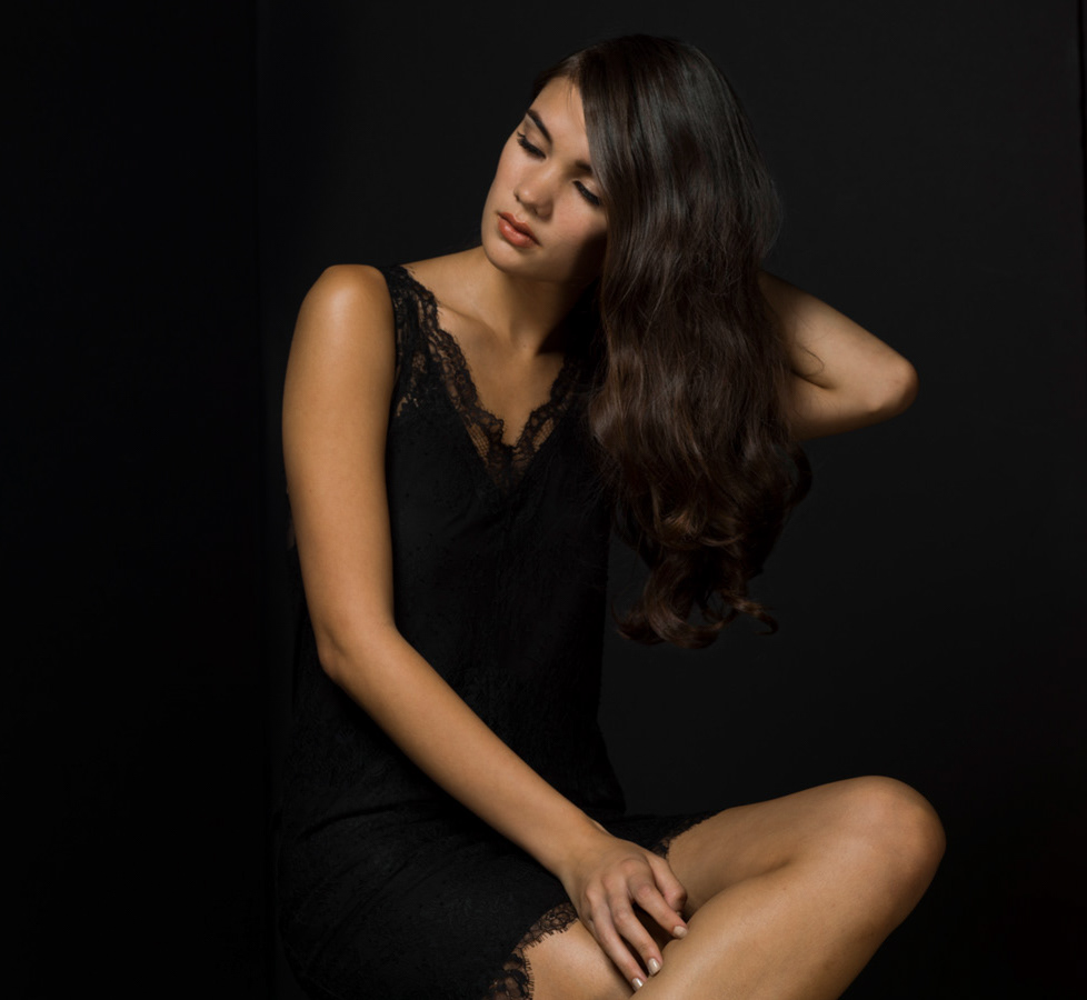 woman-black-slip-dress-legs-crossed-one-hand-on-leg-head-looking-up-and-to-the-side-with-black-background