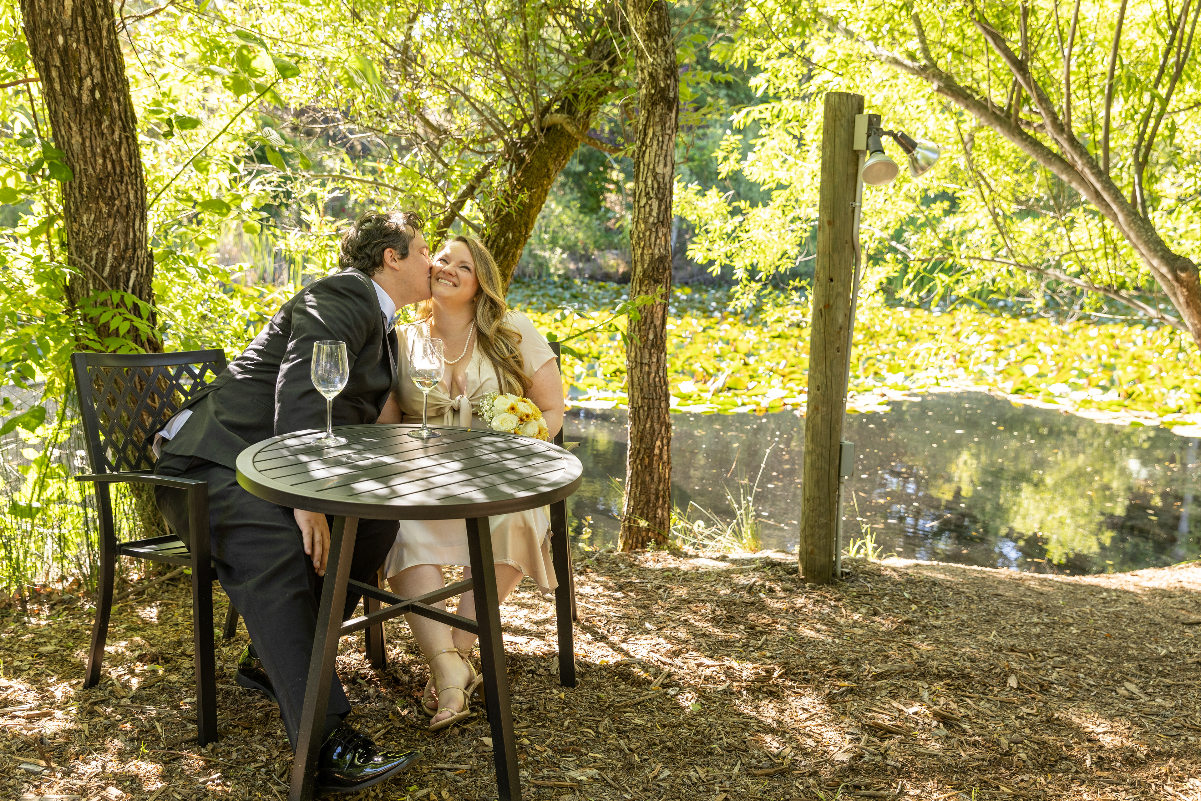 a-just-married-couple-sit-next-to-a-lily-pond-the-groom-kissing-the-smiling-brides-cheek