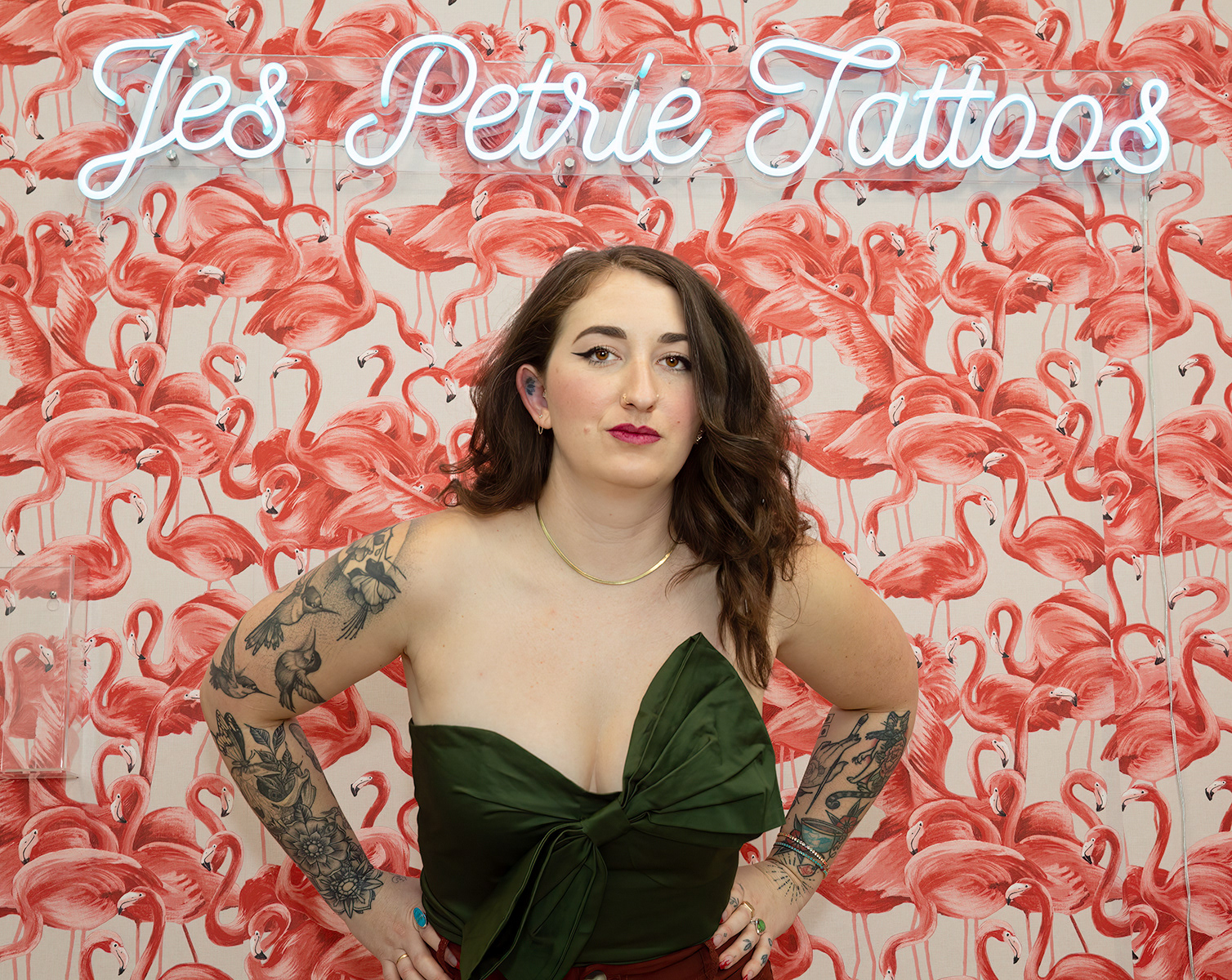 Tattooed-woman-in-strapless-green-top-and-hands-on-hips-with-pink-flamingo-wallpaper-background-and neon-sign-reads-Jes-Petrie-Tattoos