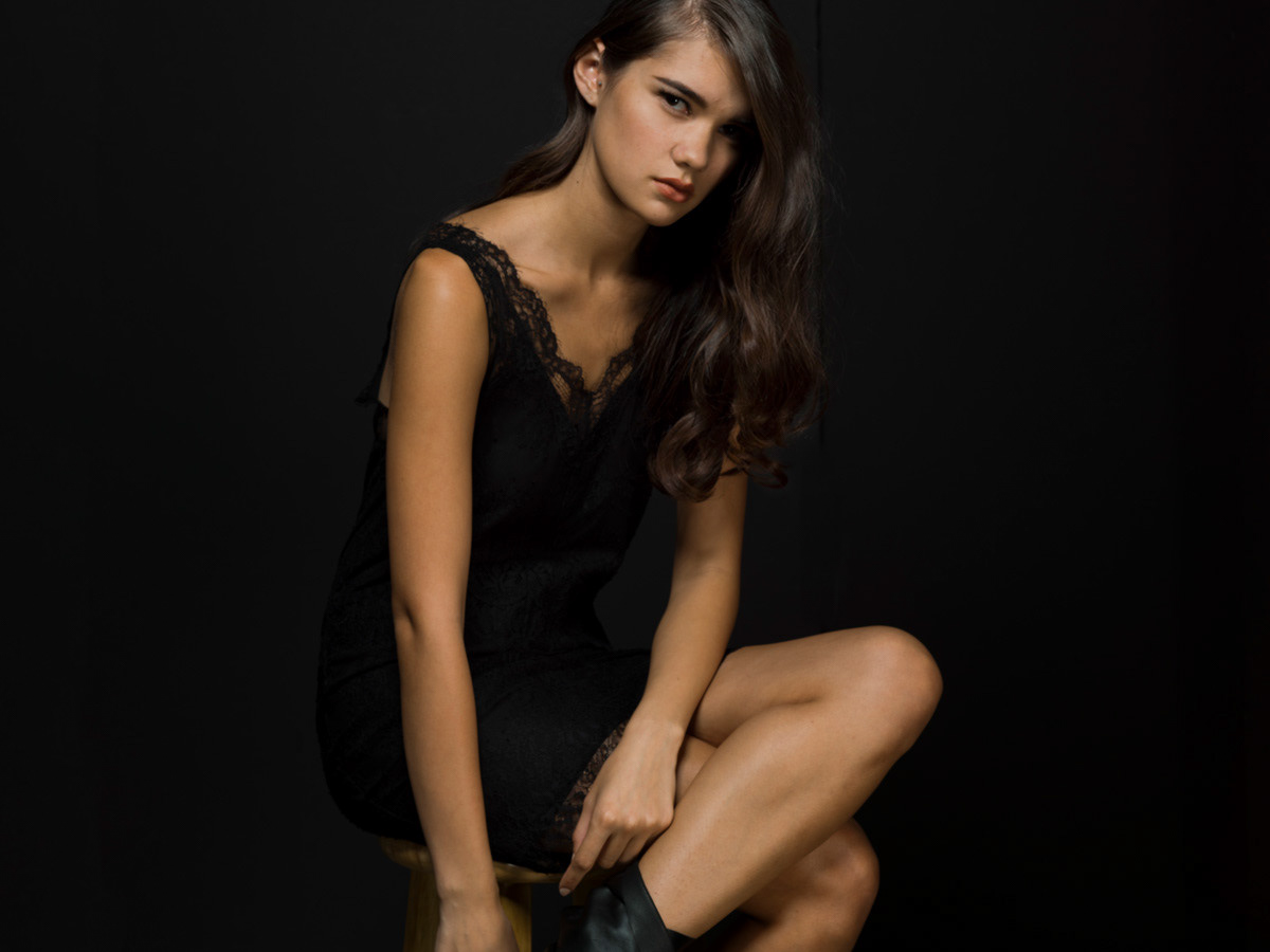 woman-black-slip-dress-legs-crossed-head-cocked-to the-side-with-black-background