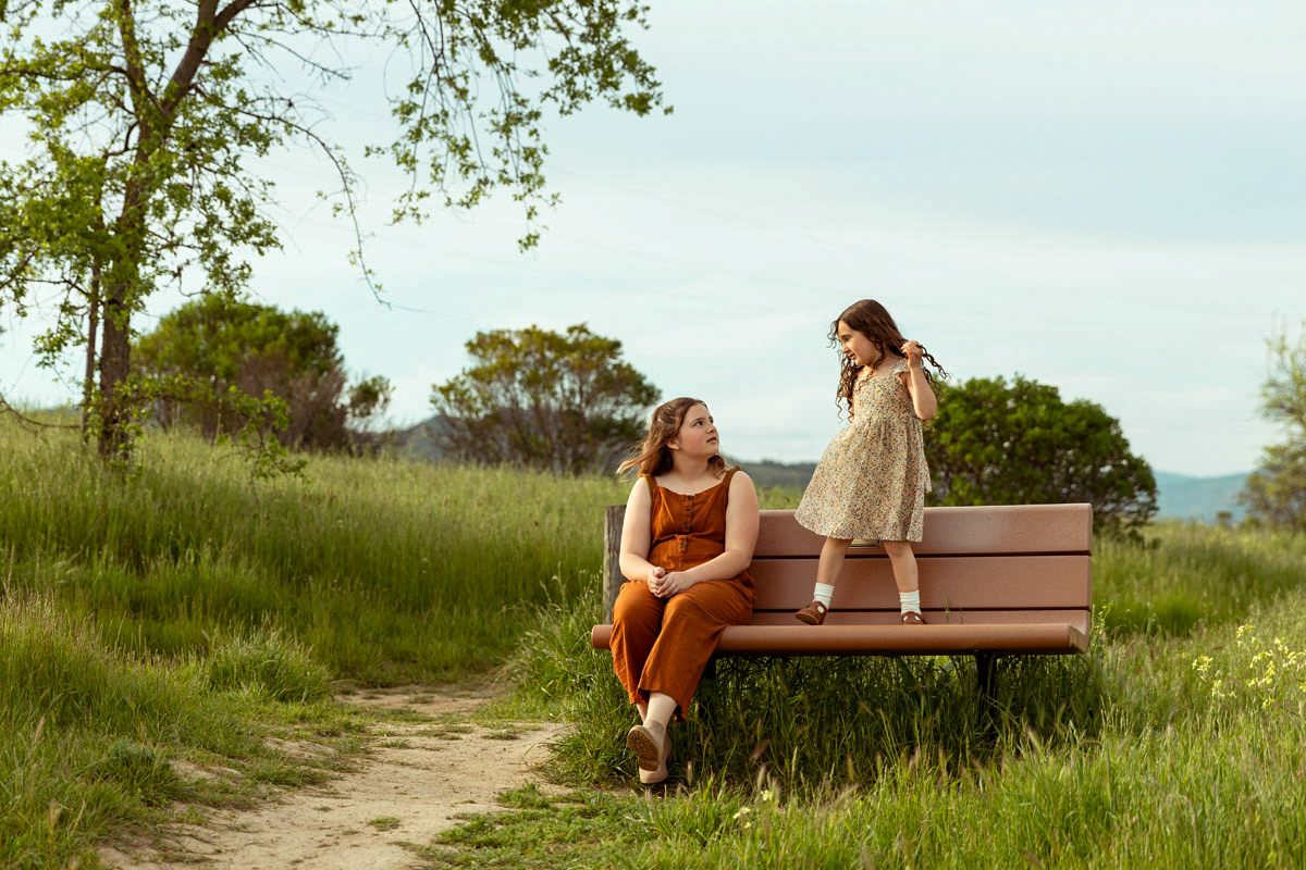 one-girls-sits-one-girl-stands-as-they-look-at-each-other-on-park-bench