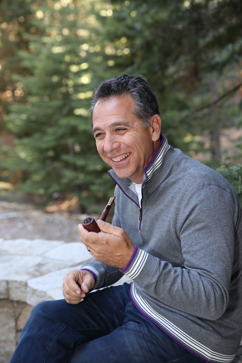 graying-dark-haired-man-smiles-and-holds-a-pipe-with-pine-trees-behind