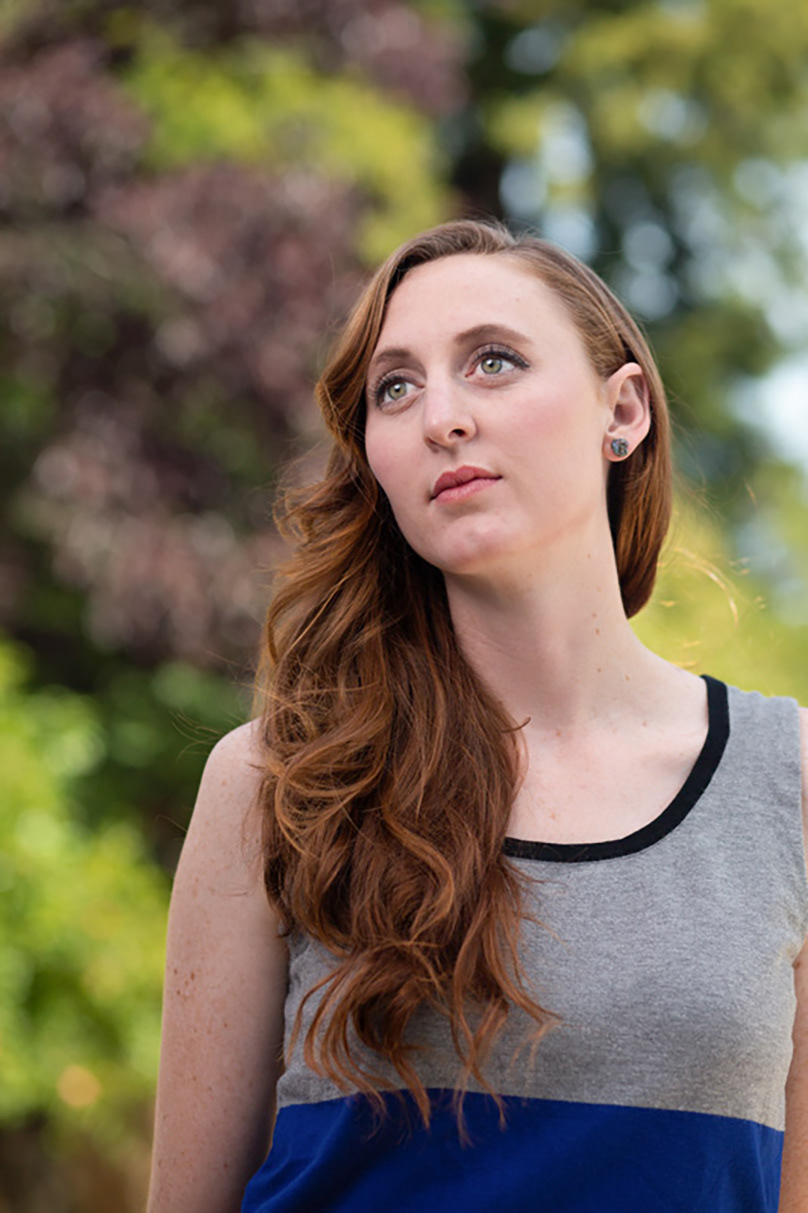 young-woman-with-green-eyes-and-long-wavy-red-hair-over-one-shoulder-wears-a-grey-tanktop-in-front-of-green-and-wine-red-folliage