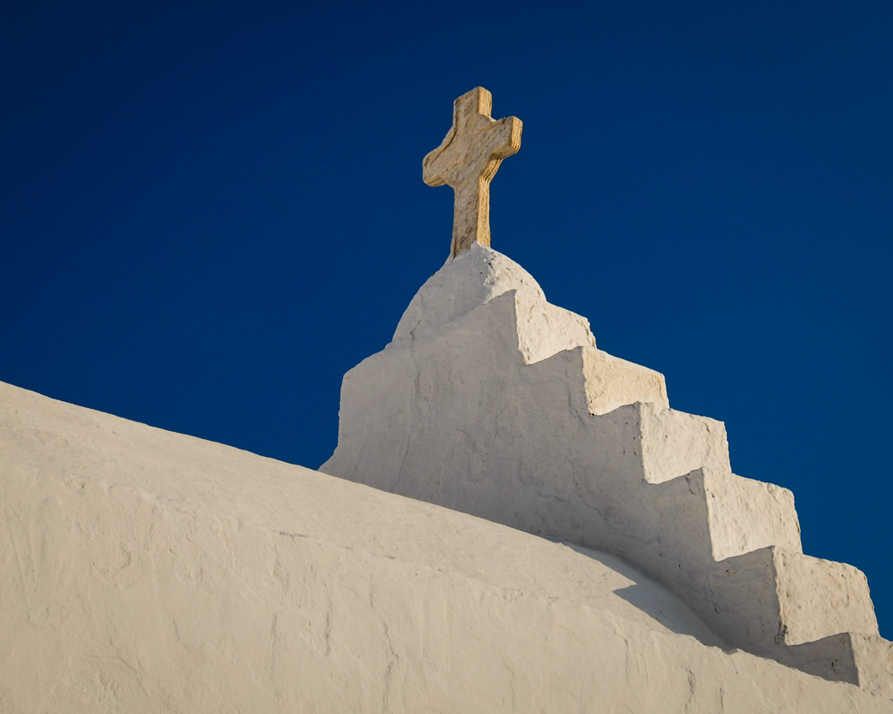 The sunlit cross sits atop the simple staircase facade of Cycladic church in Mykonos.