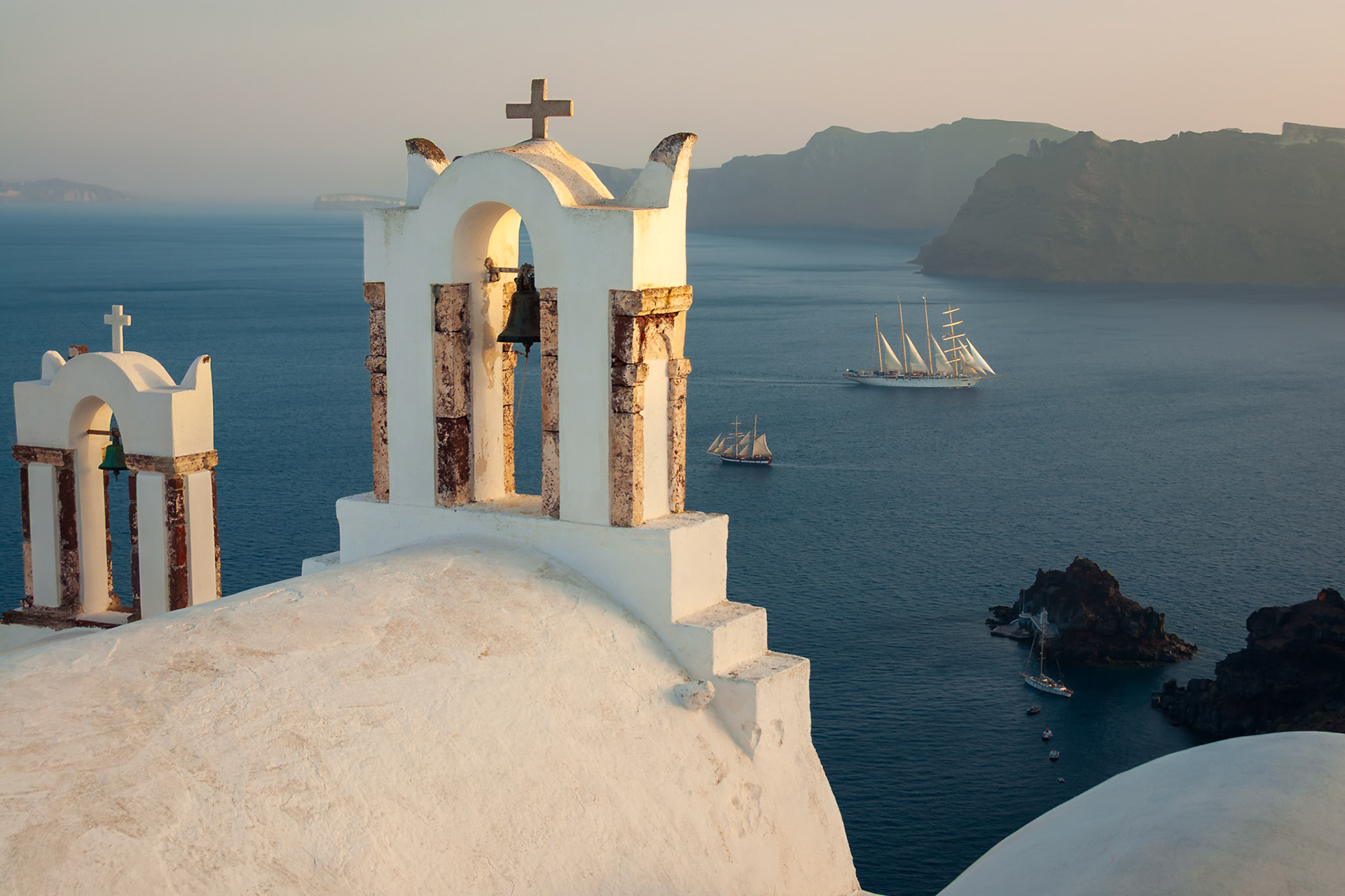 Bell towers in Oia catch the warm colors of sunset, while sailing ships cruise past Santorini's volcanic caldera.