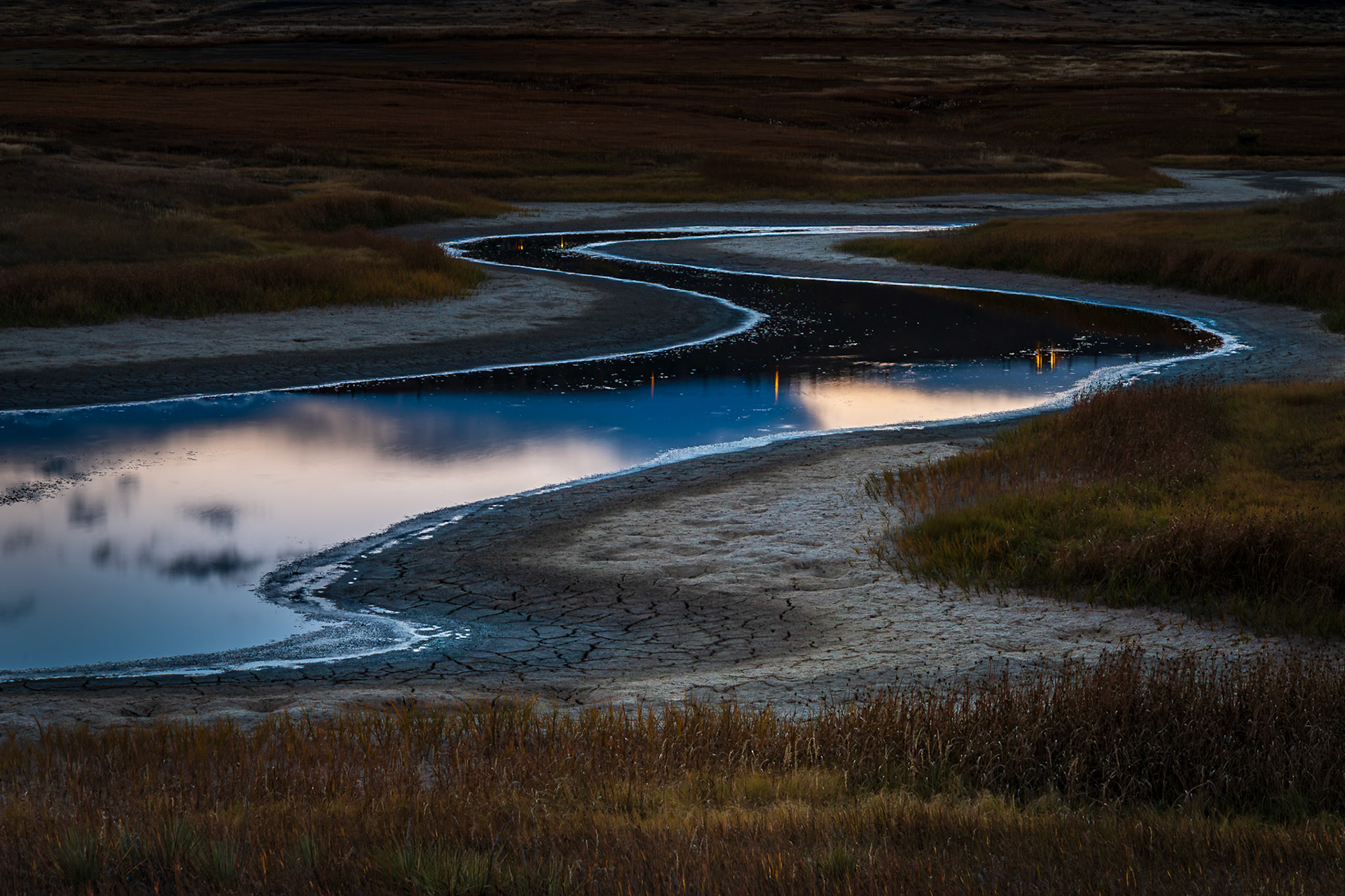 A winding stream, cradled by dry marsh grasses, mirrors the sky's pastel colors in the calm of dusk.