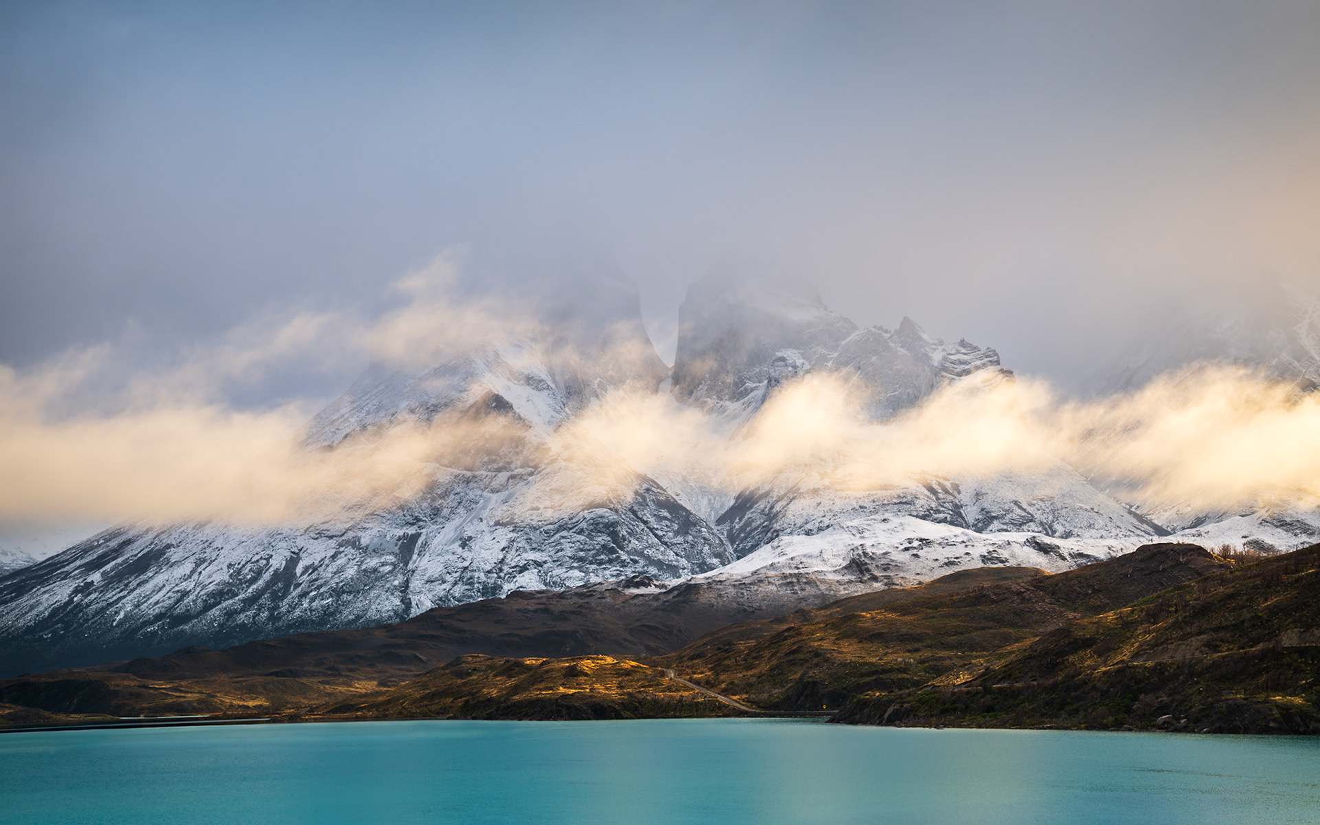 As clouds part briefly, the snowy peaks of Torres del Paine emerge over Lago Pehoé in soft, diffused morning light.