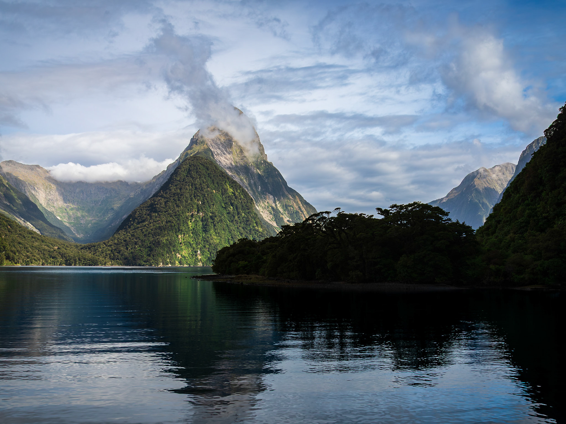 Cloud wisps swirl around Mitre Peak as it rises above the calm surface of Milford Sound.
