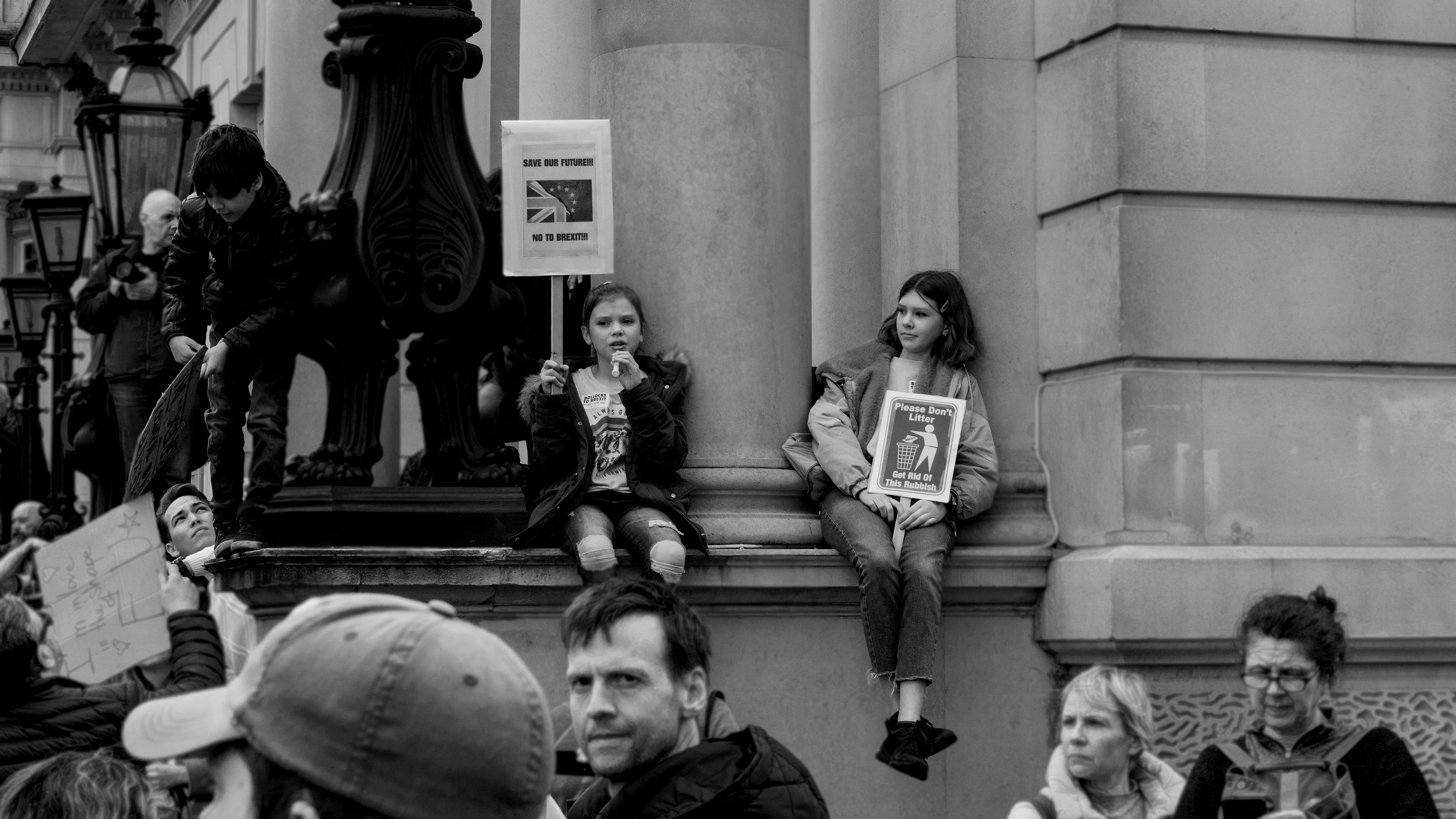 Black and white documentary photography captured in London, UK during the Brexit March 2019. Two young girls are sitting above other people. They do not look happy, also, they are holding posters against Brexit. 