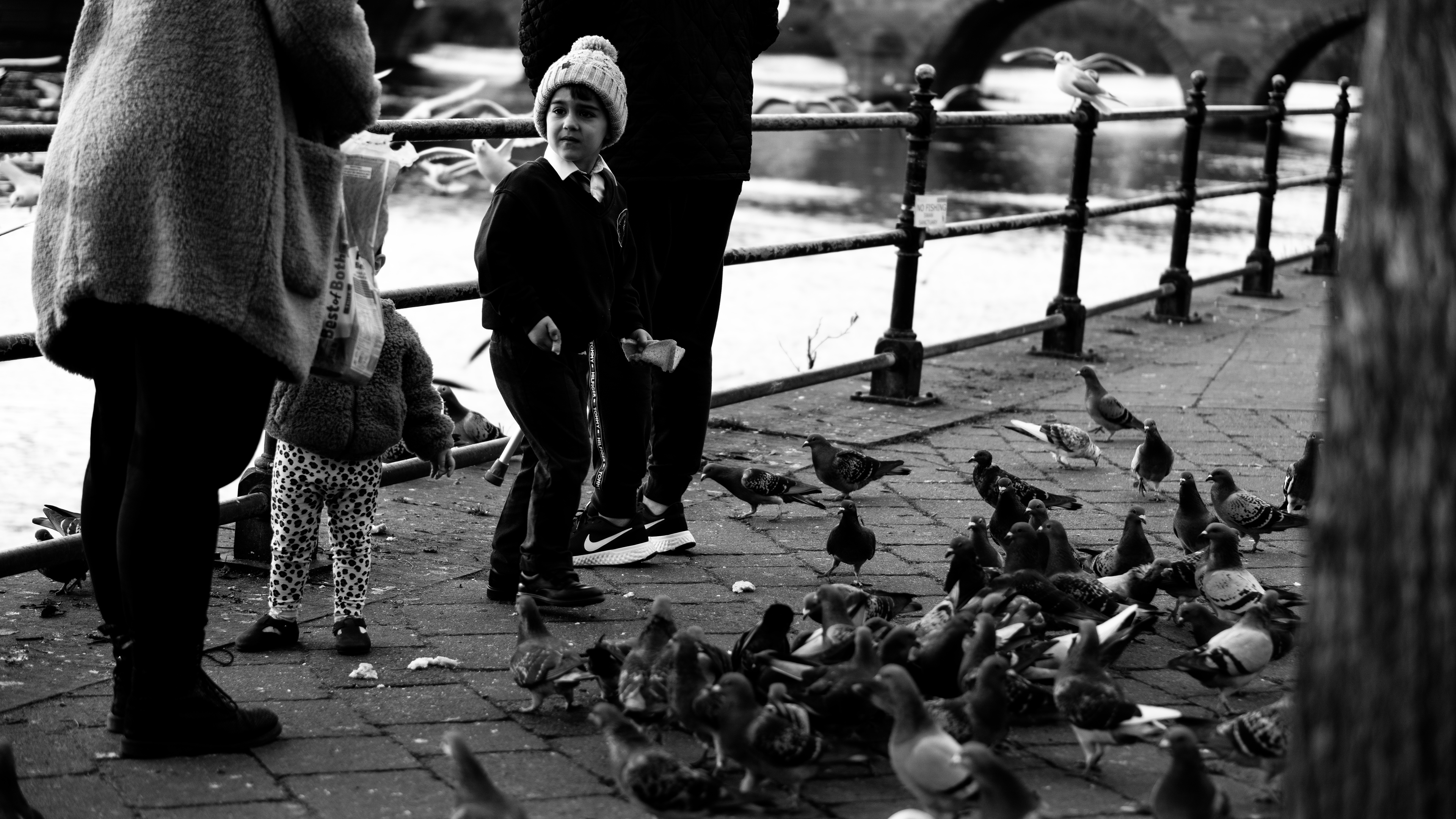 Black and white street photography. In the image there is captured a young boy with his mother, he is feeding birds