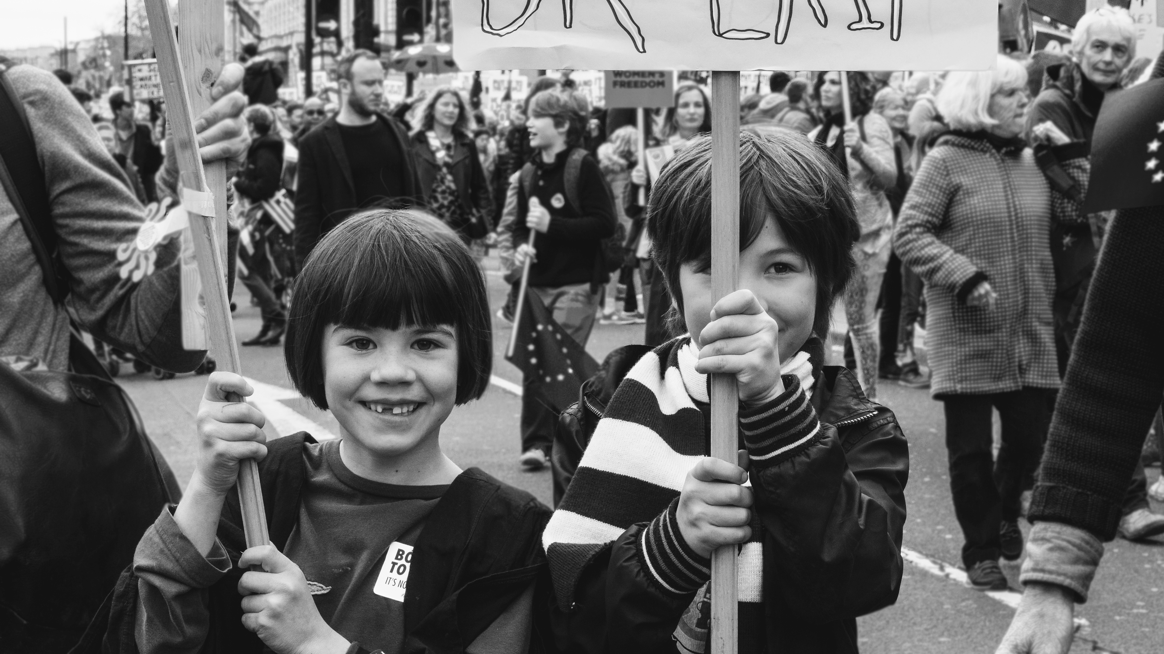 Brexit march, 2019, London, UK. This event | street photography captured two kids holding the massive poster agains the Brexit. Black and white portrait photography gives a sense of past. Brexit is like a big step back. This photograph remind of horror story, because of one of the children haircut and smile without one tooth. 