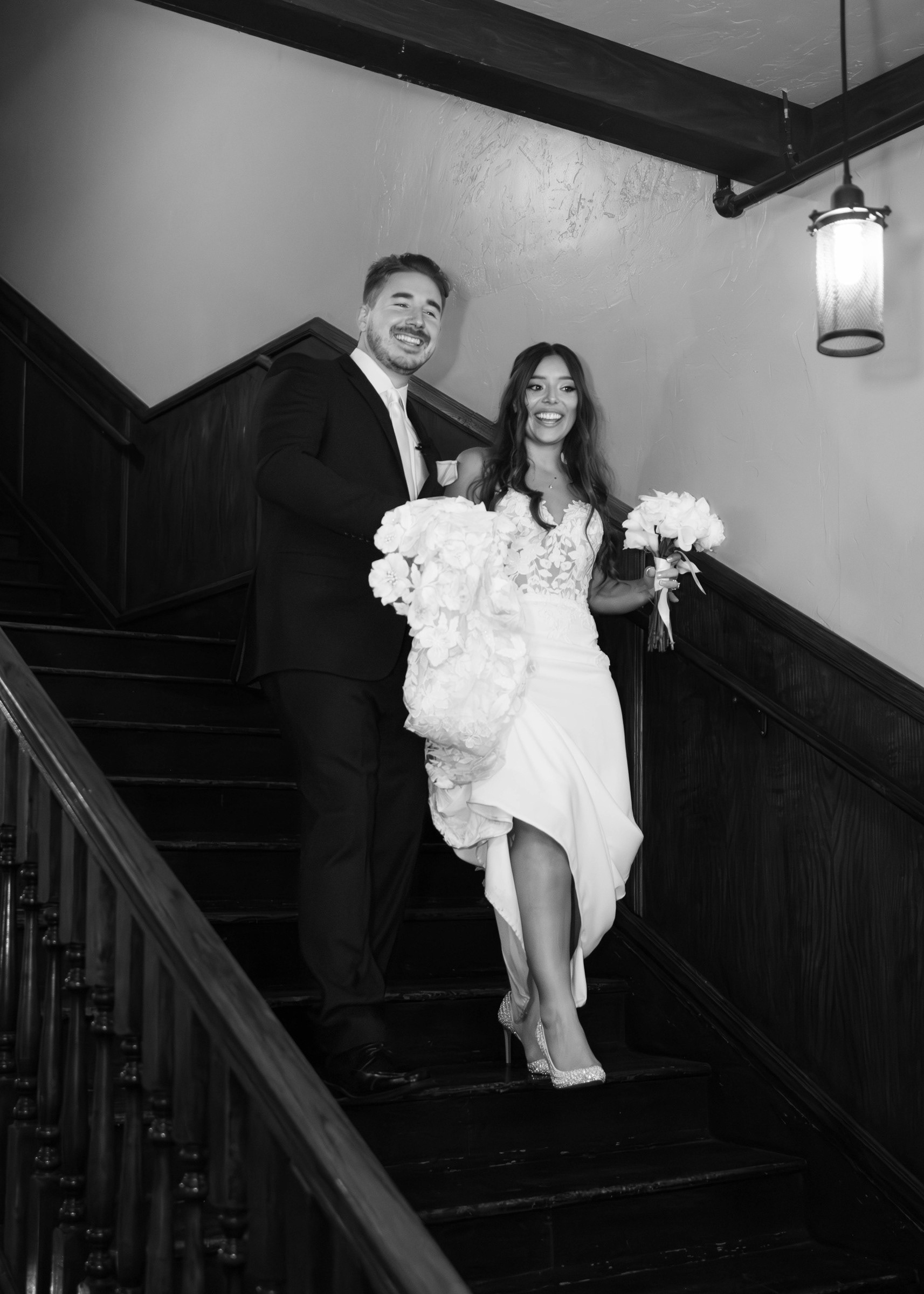 A black-and-white wedding portrait of the Bezanskys on a sweeping wooden staircase, with the bride holding her bouquet.