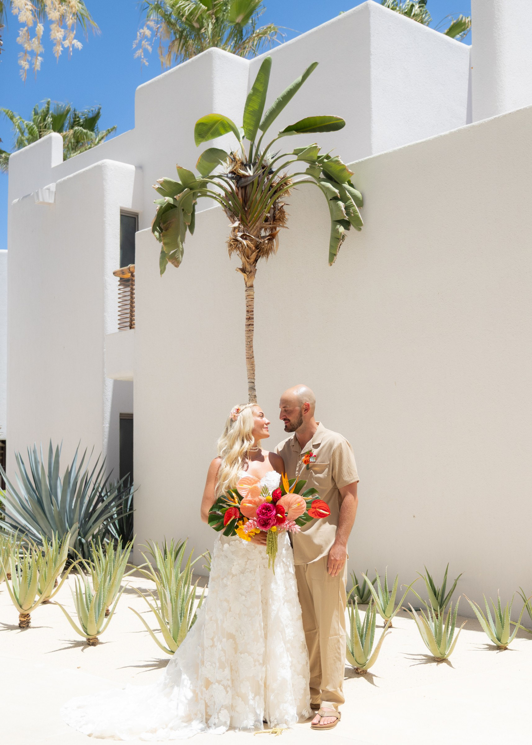 The Tuthills together beside white architecture and palm trees, holding a colorful wedding bouquet.